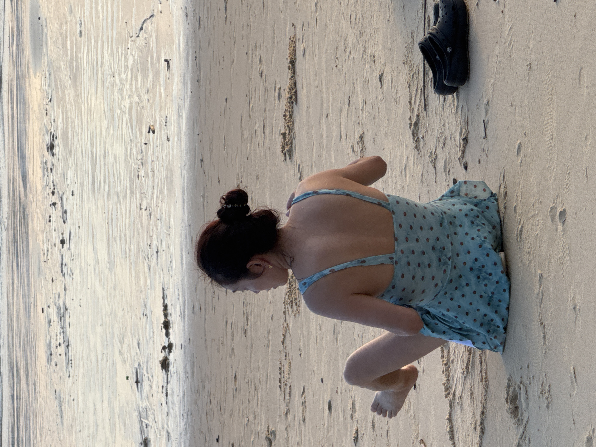 A woman, viewed from the back, sits on a sandy beach in Karon, Thailand. She has her dark hair pulled up in a bun and is wearing a light blue dress with red polka dots. Her left leg is bent, and her foot is visible. To her right, a pair of black Croc-style shoes are placed on the sand. The beach is wet in the foreground, indicating the tide is receding, and the ocean stretches out in the background. The sand shows patterns of water ripples and some debris.