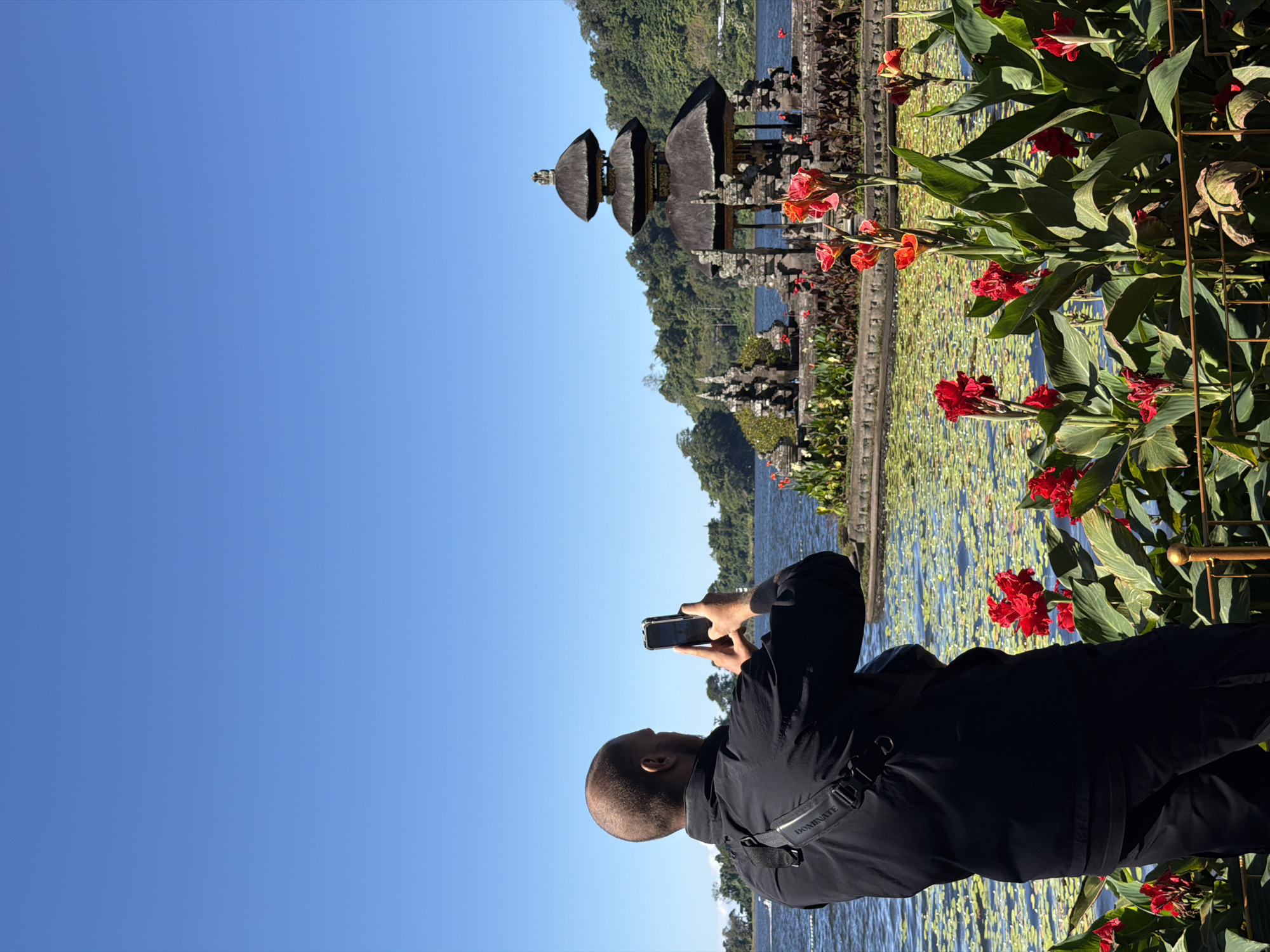 A man in a black jacket and pants is standing in the foreground, taking a photo of a temple with his phone. He is bald and has a bag slung across his chest. In the background, there is a body of water covered in lily pads, and a temple complex with multiple tiered roofs. The temple is surrounded by lush green trees. The sky is a clear blue. This is likely the Ulun Danu Beratan Temple, located in Baturiti, Indonesia.