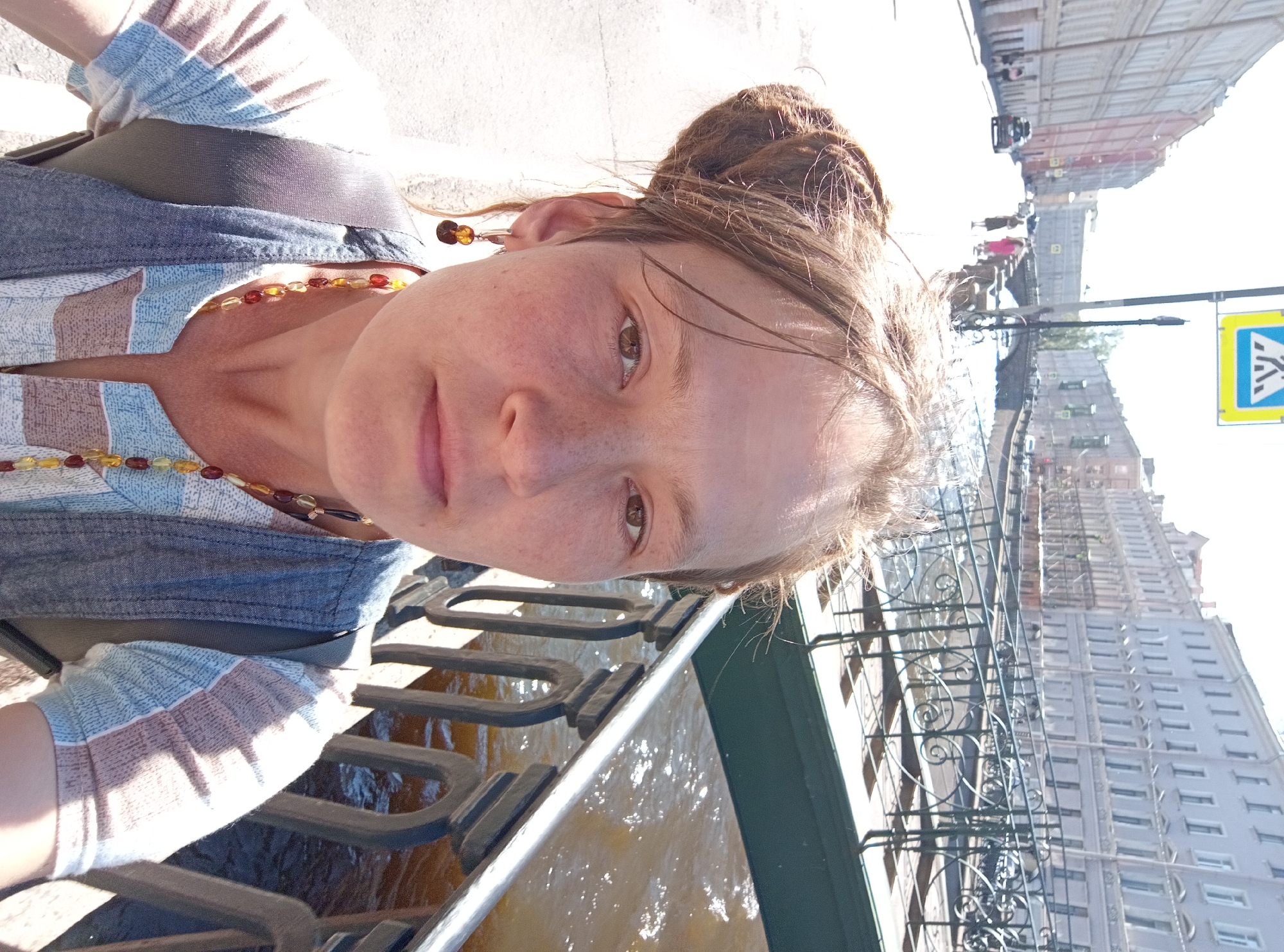 In Saint Petersburg, Russia, a woman with dreadlocks pulled up into a bun takes a selfie on a sunny day. She is standing on a street with tall, old buildings lining one side and a canal with a decorative black iron bridge and railings on the other. The canal water is a muddy brown. The woman wears a blue denim vest over a striped shirt, a beaded necklace, and amber earrings. In the background, a pedestrian crossing sign is visible, and several people are walking along the street and the canal. Cars are also visible on the street. The image appears to have been taken midday due to the bright sunlight and harsh shadows.