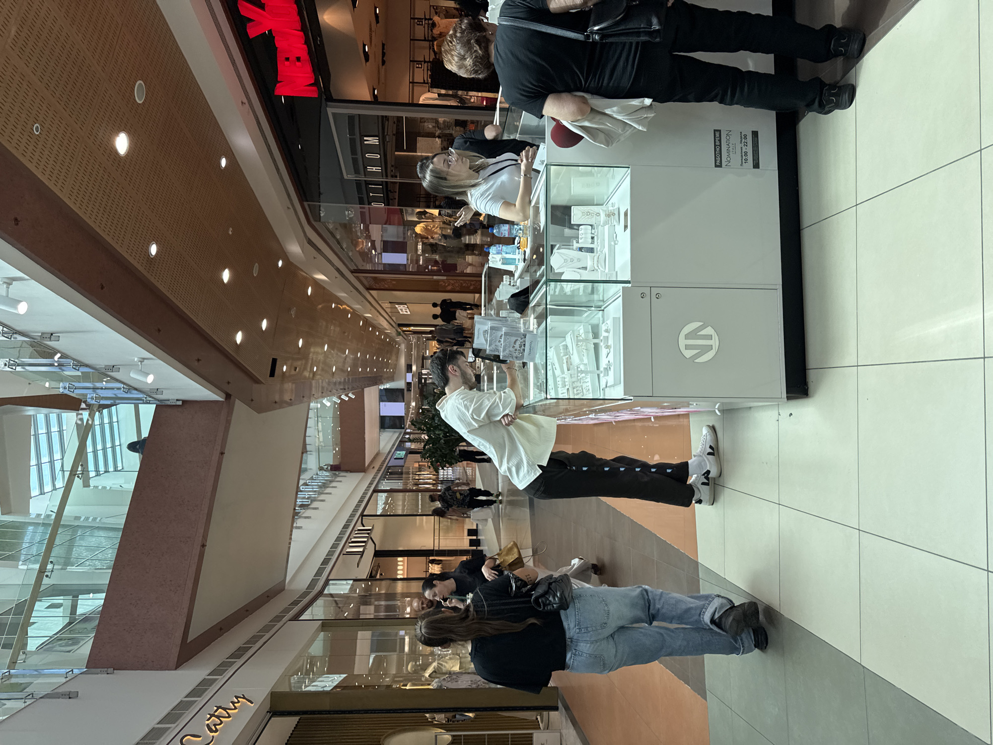 The image shows an interior view of what appears to be a shopping mall. At the front of the view is a jewelry store, "NOMINATION Italy," with glass display cases showing items. A store employee stands behind the counter, attending to customers. Several people are looking at the jewelry. The mall is well-lit with a variety of shops visible in the background and people walking around.
Based on the store name, the location may be in Sofia, Bulgaria. The store's operating hours of 10:00-22:00 are displayed.