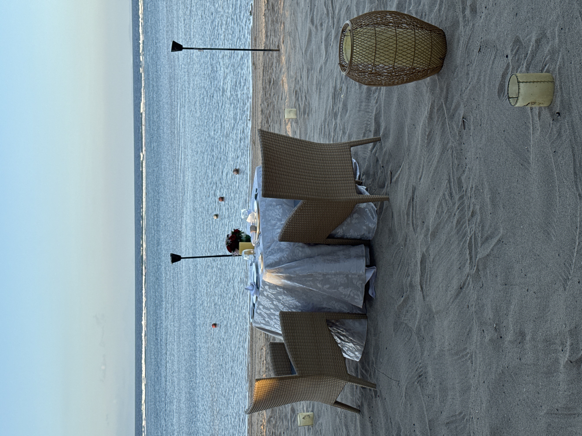 A romantic dining setup for two is arranged on a sandy beach in Kuta Selatan, Indonesia, overlooking the ocean. Two wicker chairs face a circular table draped with a light-colored, patterned tablecloth. The table is set with plates, silverware, glasses, and a vase of red flowers. Two tiki torches stand behind the table, and several small, cylindrical candle holders are placed in the sand around the dining area. A large, woven lantern sits to the right of the table. The calm ocean stretches to the horizon under a clear blue sky.