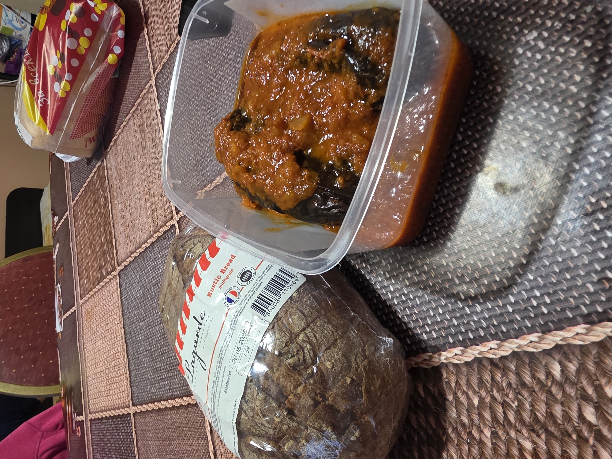 Here is a description of the image:
The image depicts a table setting with food items. A clear plastic container holds a dark-colored food, possibly eggplant, covered in a chunky orange sauce. Next to it is a loaf of dark, crusty bread in a plastic wrapper. The bread label is partially visible, revealing the brand name "Lagarde Rustic Bread multigrain" and a best before date of "26 05 2026". A bag of bread with a colorful design is placed to the left of the container. The table itself has a textured woven surface in brown and beige tones. Part of a red chair is visible on the left side. The location cannot be determined as Yagodovo, Bulgaria from this information.