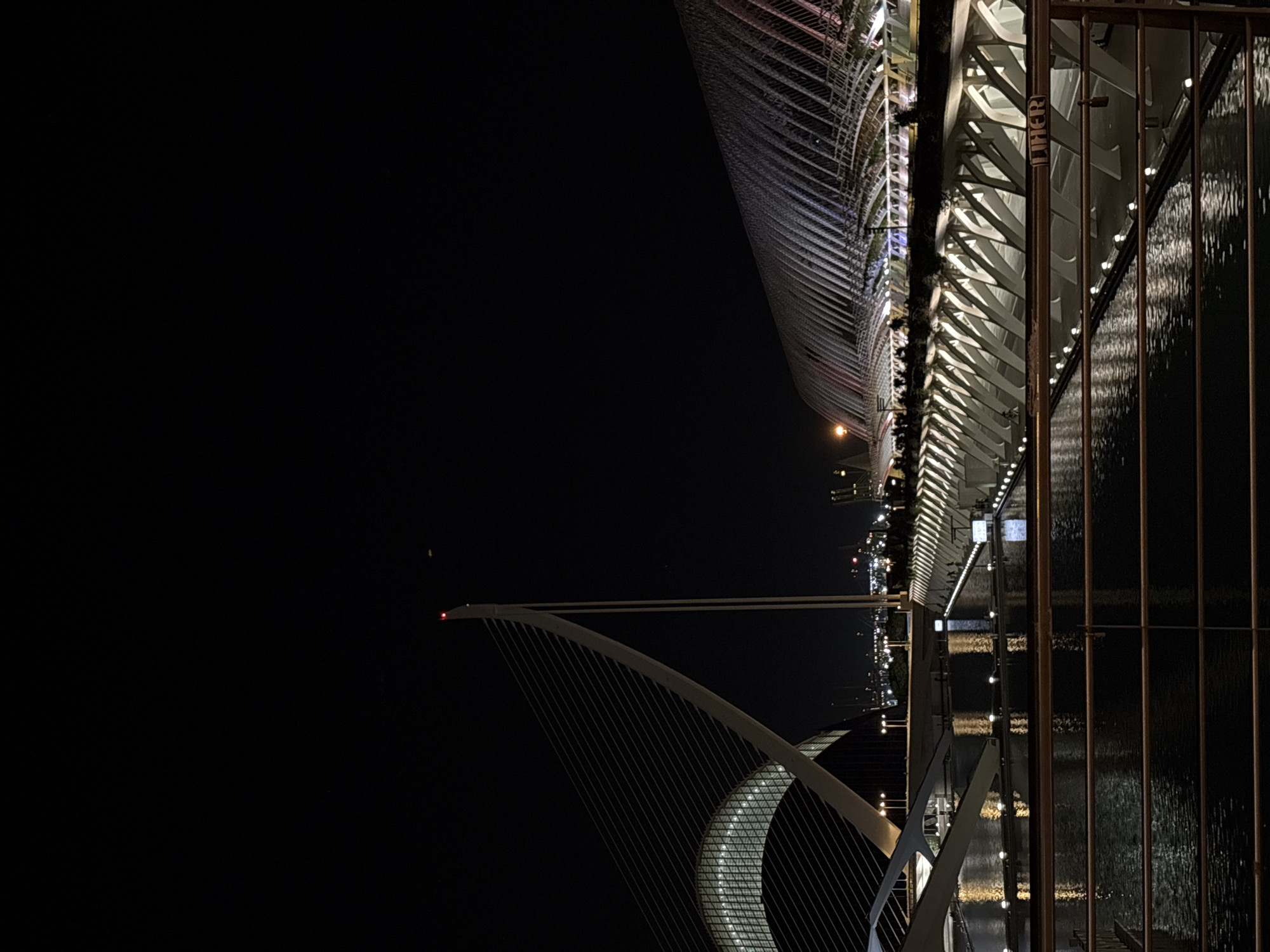 The image captures a nighttime view of the City of Arts and Sciences in Valencia, Spain. The foreground features a dark body of water reflecting the lights of the surrounding architecture and a metallic railing with a lock attached. Dominating the scene are two distinct modern architectural structures. On the left, a cable-stayed bridge with a tall, slender pylon topped with a red light is prominent. Behind it, a large, arched building with a translucent roof is partially visible. On the right, a long, curved structure with a distinctive ribbed roof, illuminated with multi-colored lights, stretches across the frame. The water in front of the structures is calm, with numerous small lights lining its edges and reflecting the vibrant illumination of the buildings. The sky is dark, indicating nighttime. There are no discernible people or actions depicted.