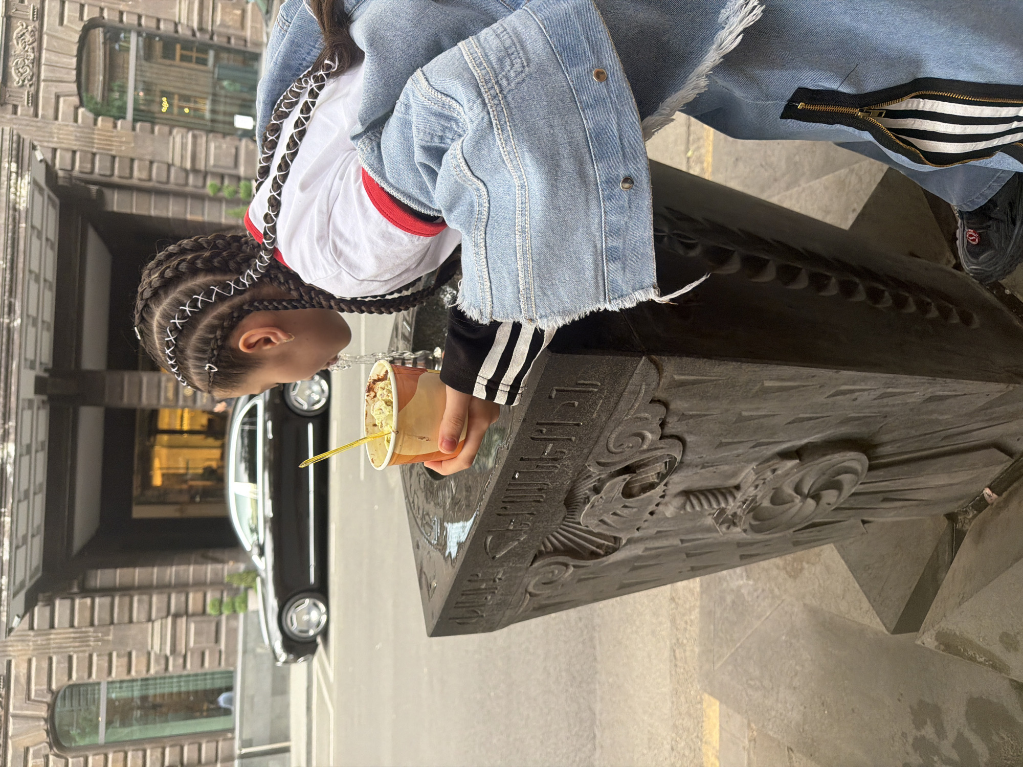 A young girl with braided hair stands in Yerevan, Armenia, next to a large, gray stone monument. The girl is bending forward, appearing to eat something from a cup with a yellow straw. She wears a white shirt with red trim, a denim jacket, and black athletic shorts with white stripes. The monument has decorative carvings and some inscriptions that are difficult to read. Behind the girl and the monument is a black car parked on the street and the entrance to a building with a decorated facade. The scene is outdoors during daylight.
