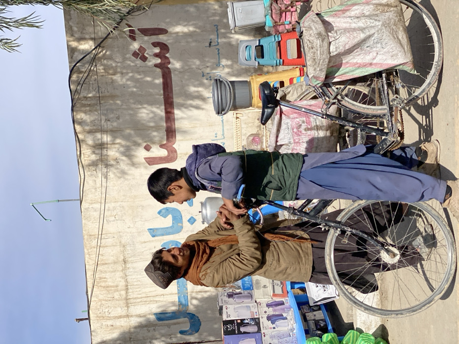 This image captures an outdoor scene in Kandahar, Afghanistan, under bright sunlight, depicting two males interacting near a bicycle. An adult male, positioned on the left, wears a brown jacket, a patterned brown scarf, and a dark embroidered cap. He faces right with a pleasant expression, extending his hands to interact with the hands of a younger male. The younger male, standing on the right, wears a blue shalwar kameez and a dark green vest. He holds the handlebars of a black, cargo-modified bicycle equipped with large, light-colored sacks on both its front and rear racks.
Behind the individuals is a weathered, light-colored concrete wall featuring painted script: blue characters resembling "بابا" are on the left, and prominent red characters spelling "آشنا" are on the right. To the adult male's left, a blue display shelf unit holds multiple thermos flasks. To the younger male's right, various household items are stacked, including a grey bucket on a yellow plastic stool, two additional colorful plastic stools, and a silver-colored metal container. Black electrical wires span horizontally across the upper wall, and a green metal pipe structure ascends into the clear blue sky in the upper left background. Partial palm fronds are visible on the far right.