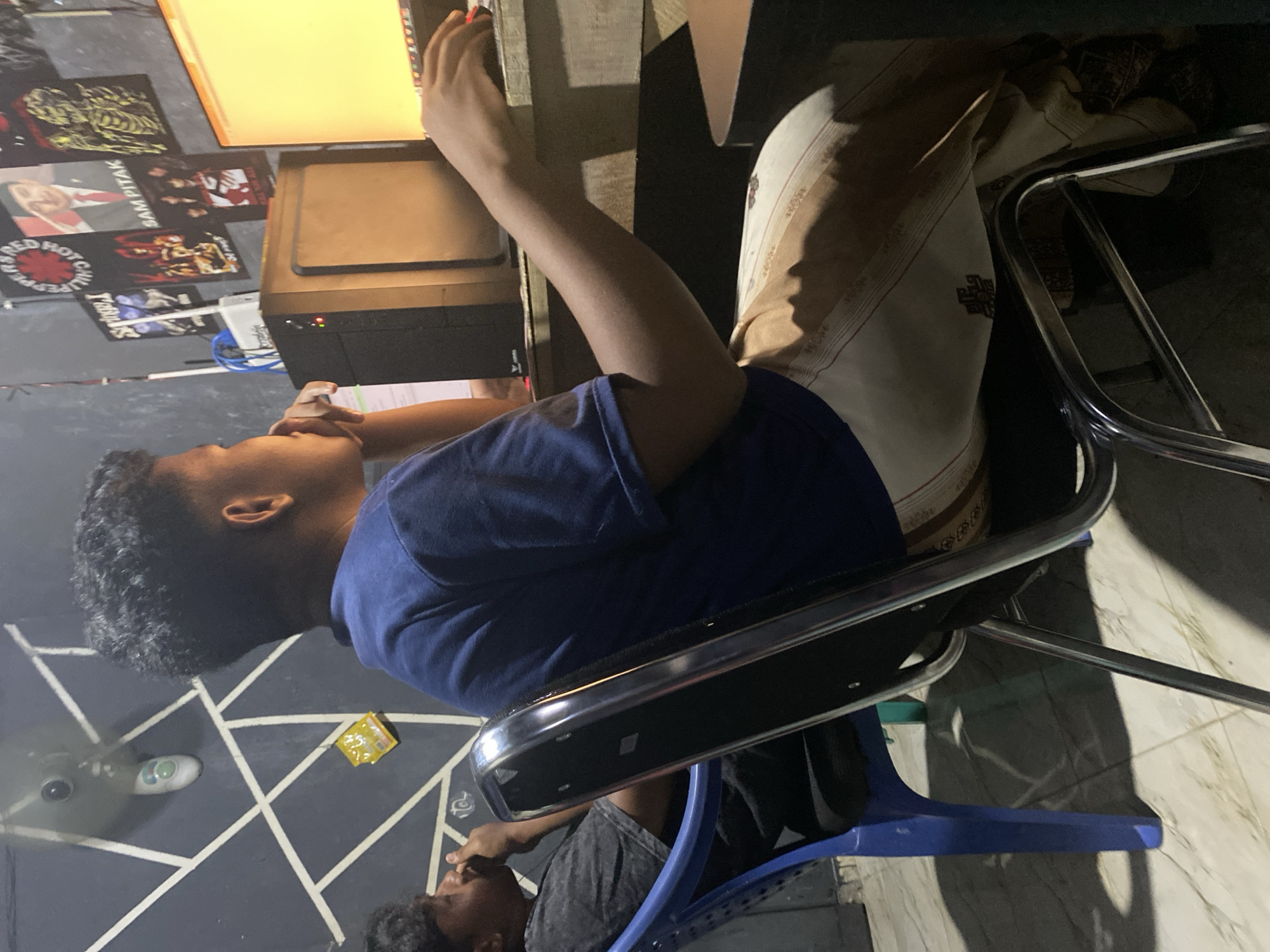 The image shows two young men in a dimly lit room, likely a home office or gaming area, in Sumber Baru, Indonesia. The man in the foreground, wearing a blue t-shirt and patterned pants, is seated in a metallic chair and focused on a computer monitor. His right hand is on the mouse. To his left, another young man sits in a blue plastic chair, wearing a dark t-shirt and appearing to be interacting with something near his face.
The room's background is adorned with geometric white lines on a dark wall. A white ceiling fan is visible in the upper left corner, along with a small yellow packet. Several framed posters, including what appears to be a Red Hot Chili Peppers poster and a portrait of a man identified as "SAM PITAK," are hung on the wall behind the computer setup. A desktop computer tower, a router with visible antennas, and a brightly lit computer monitor are placed on a wooden desk. The floor appears to be tiled. The overall impression is a casual, lived-in space with personal touches.