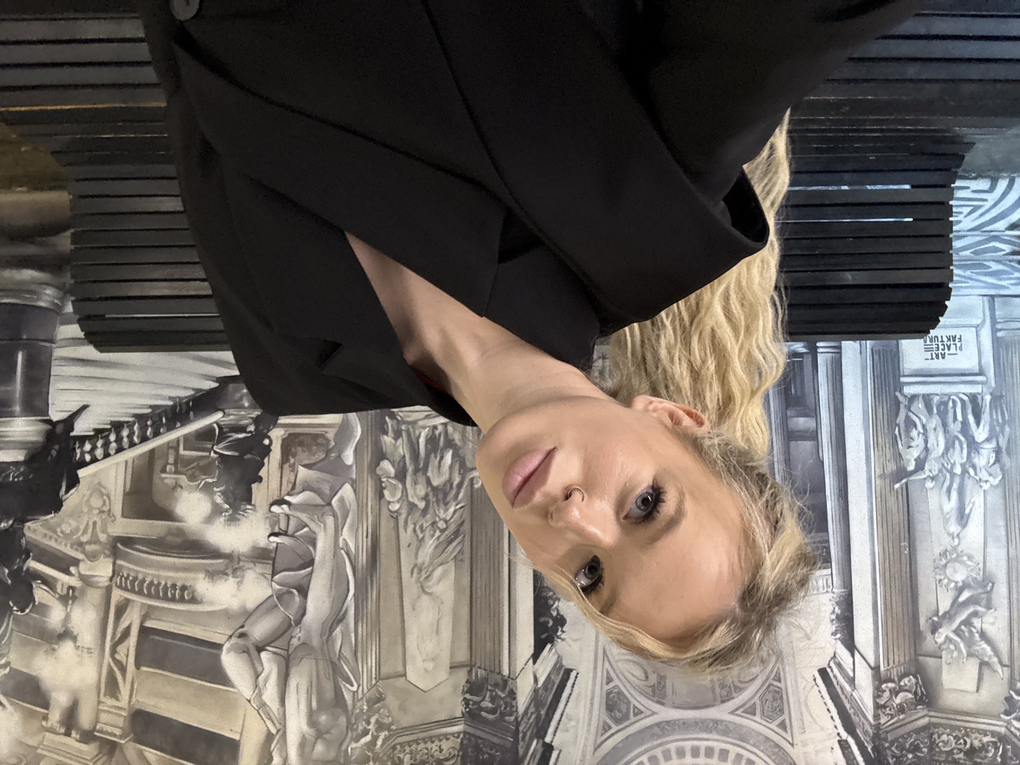 A woman with blonde hair and blue eyes is taking a selfie in front of a mural of classical architecture. She is wearing a black blazer and a nose ring. The mural depicts a grand staircase, statues, and columns. The woman is positioned slightly to the left of the frame, with her head tilted. The lighting is soft, and the overall mood is relaxed. The mural appears to be a painted scene, possibly in an indoor setting. There are also two black benches visible in the foreground, partially obscured by the woman. One bench has text on it that reads "-ART PLACE FAKTURA".