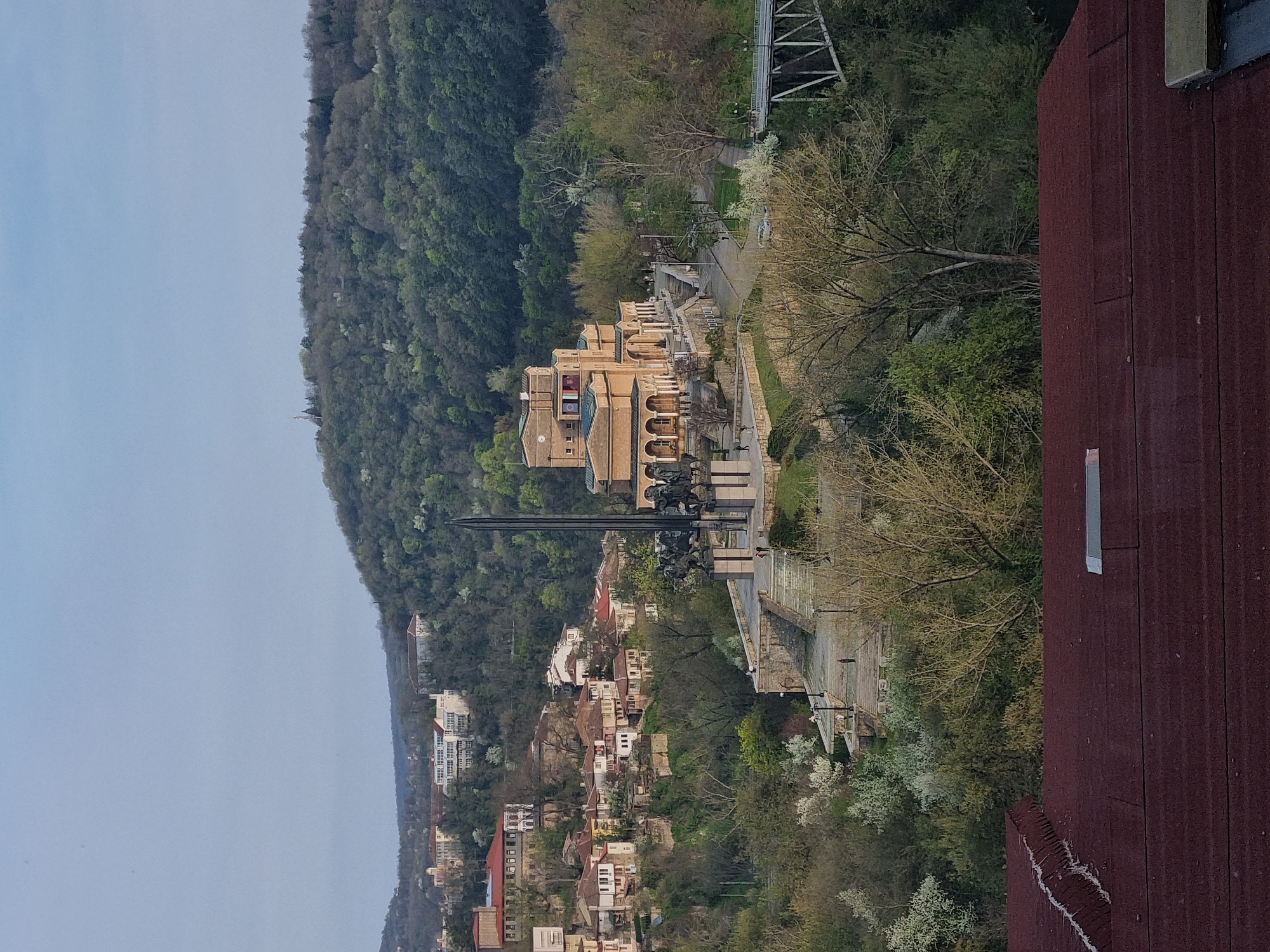The image shows a panoramic view of Veliko Tarnovo, Bulgaria, featuring the Asenevtsi Monument and a portion of the city's architecture nestled amidst green hills. The Asenevtsi Monument, with its prominent black spears, is centrally located in the view. The monument overlooks a cluster of residential buildings and winding streets below. The landscape surrounding the city includes a dense forest covering the hills in the background. A portion of a red-brown roof is visible in the foreground, suggesting the viewpoint is from a higher building. A pathway stretches parallel to the monument. A bridge-like structure is partially visible in the upper-right. The sky is overcast and a soft blueish-gray.