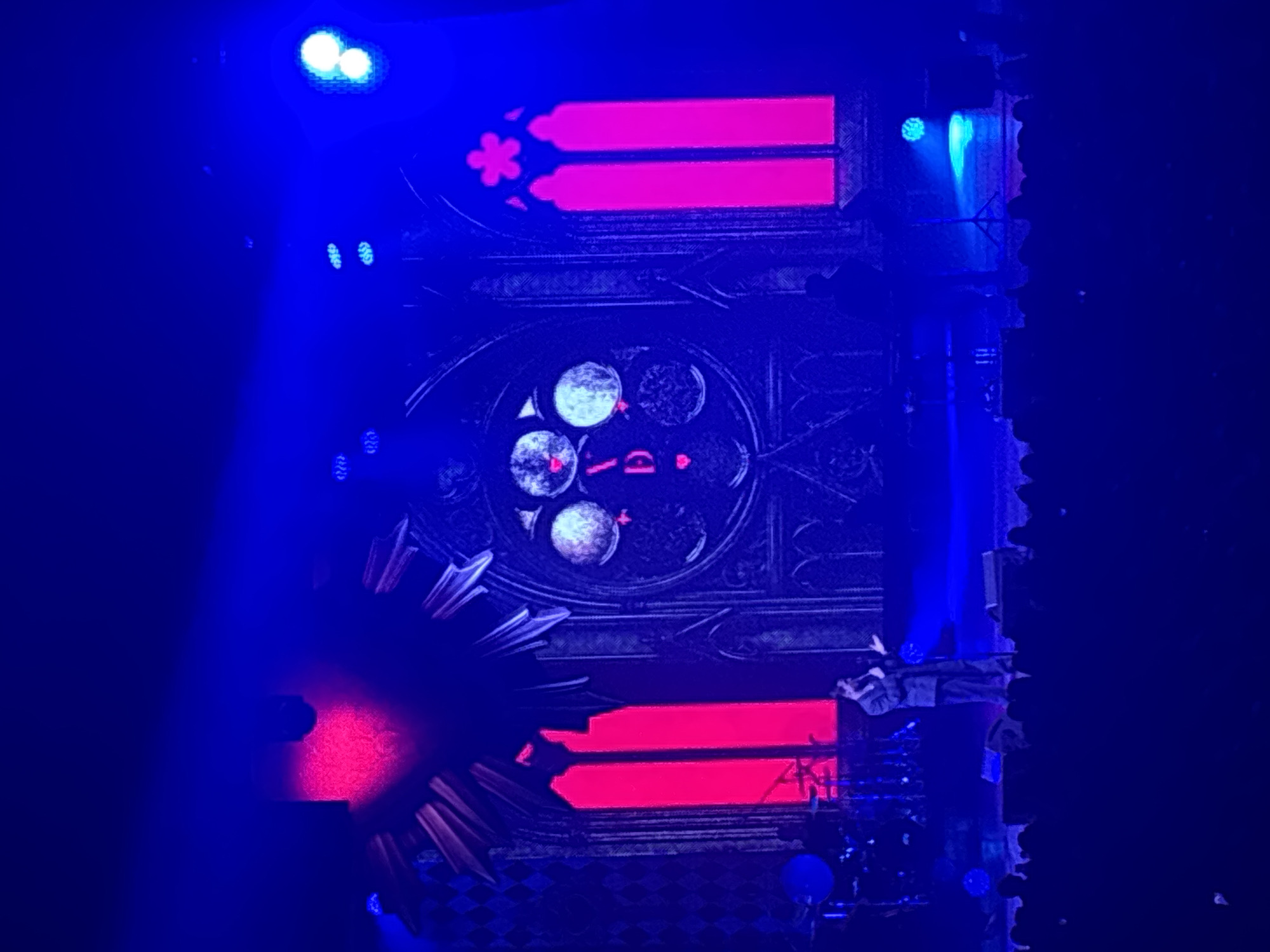 The image shows a stage set, potentially a theater or concert venue, in what may be the city of Perm, Russia. The central element is a large, dark-colored, multi-tiered structure with gothic-inspired architecture. This structure features horizontal, rectangular sections illuminated in bright red, and a circular, window-like element with illuminated white circles. The structure is detailed with patterns resembling stained glass or intricate metalwork.
Stage lighting is prominent, with strong blue beams and smaller spotlights highlighting different areas of the set. To the right, there appears to be a rocky or textured vertical element. The overall lighting is dramatic and theatrical.
The exact location within Perm cannot be determined from the image alone.