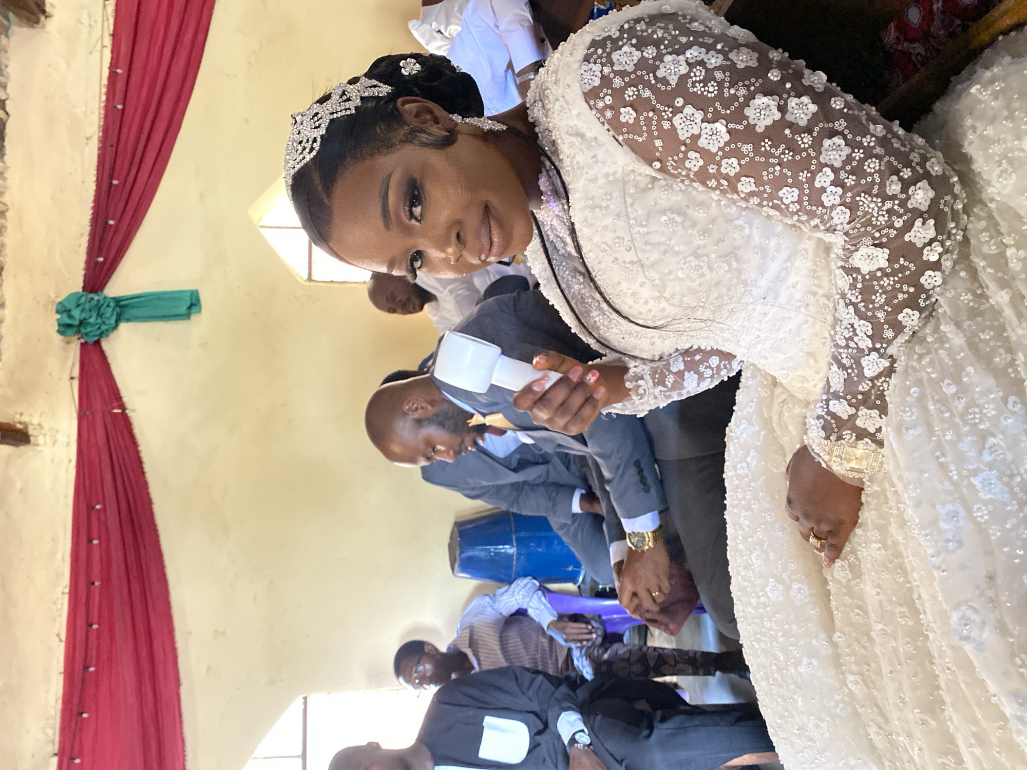 The image shows a bride indoors, likely in a church in Vom, Nigeria. She's wearing a white, long-sleeved, heavily embellished gown and a tiara. She's holding a white object, possibly a microphone or device for measurement. Several men in suits are visible in the background; they appear to be part of the wedding party or guests. Some are looking at cards or documents. A red and green decoration hangs on the wall.