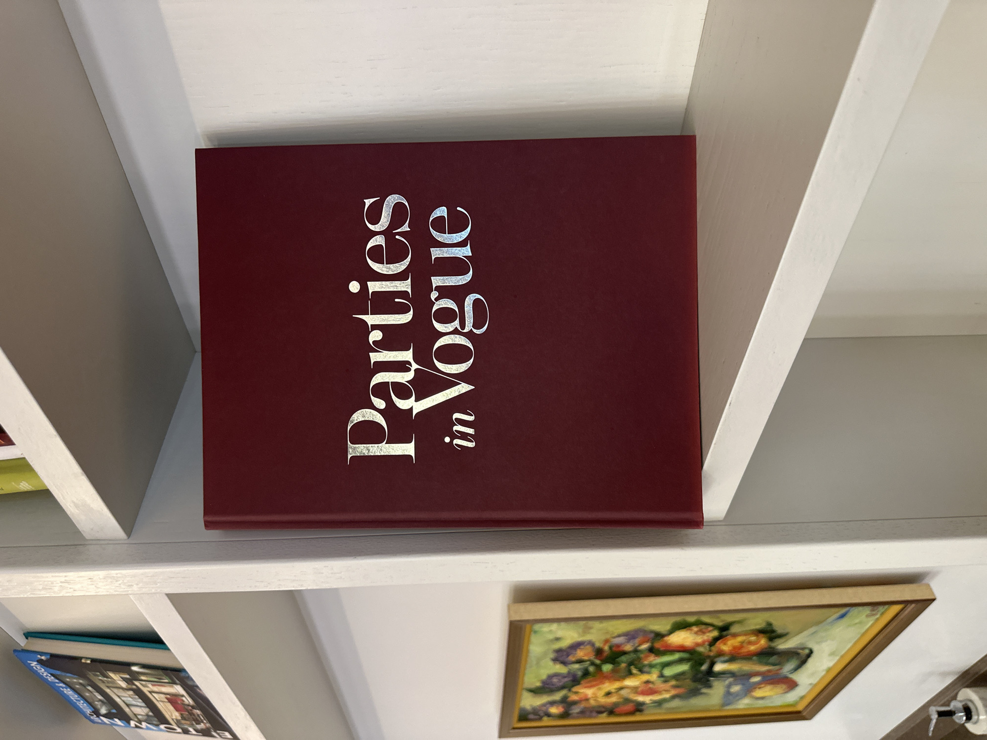 A close-up shot shows a book titled "Parties in Vogue" in silver lettering on a burgundy cover. The book is sitting on a white shelf within a white bookshelf unit. Below the book and the shelf is a framed painting of flowers. Other books are partially visible on shelves above and below. The photo suggests an indoor setting, possibly a home or bookstore. The location is not discernible from this image.