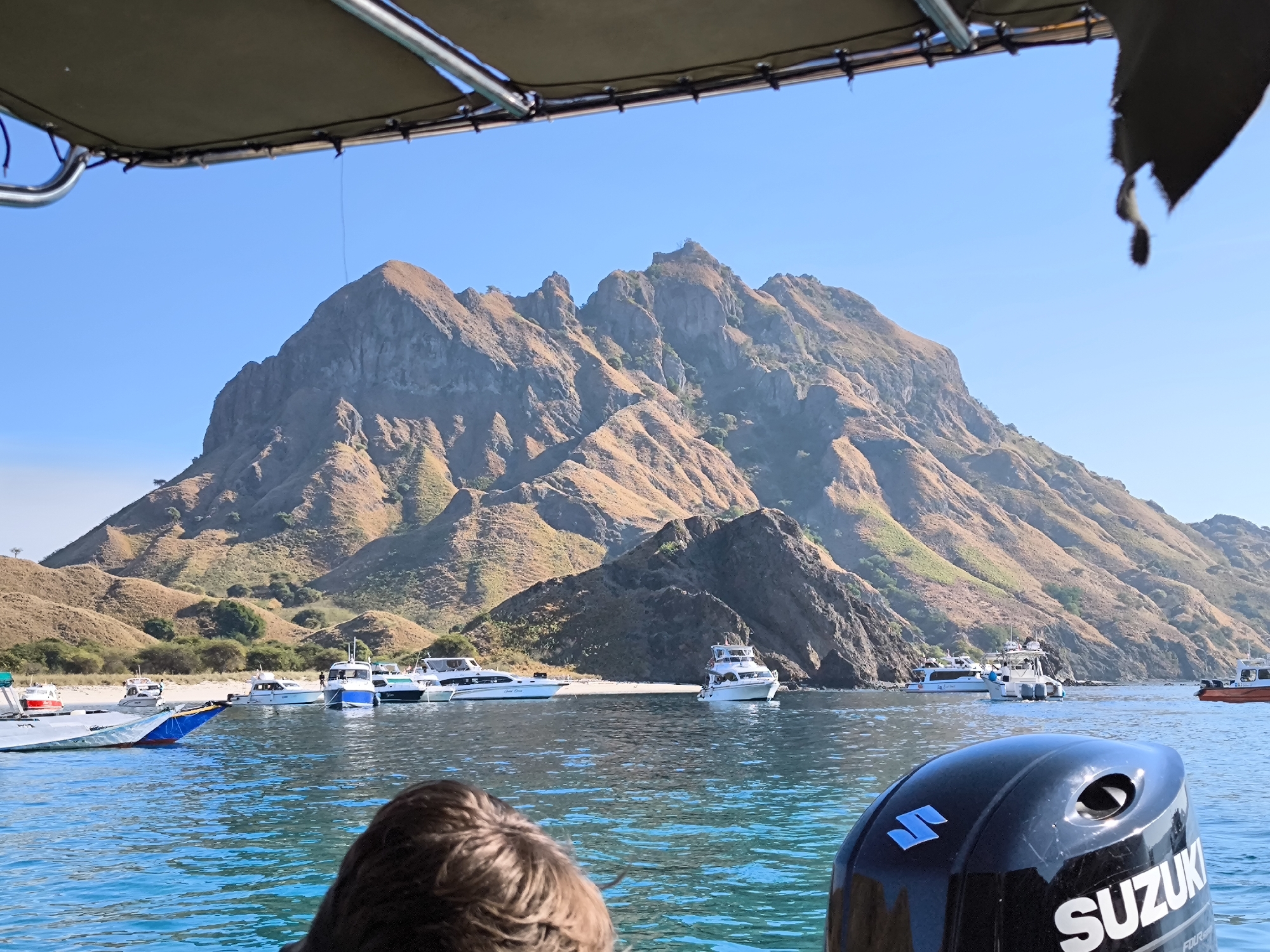 The image captures a scenic view of a mountainous island from the perspective of a boat. The foreground shows the back of a person's head and the outboard motor of a boat, with "SUZUKI" clearly visible on it. The water is a vibrant blue, reflecting the clear sky. In the middle ground, several boats of varying sizes are anchored in the water near a sandy beach. Beyond the beach, the island rises steeply, characterized by dry, golden-brown vegetation and rugged rock formations. The overall scene is one of a tranquil coastal environment in a tropical or subtropical region. The location is likely Padar Island, Komodo National Park, Indonesia, a popular destination known for its unique landscapes and clear waters.