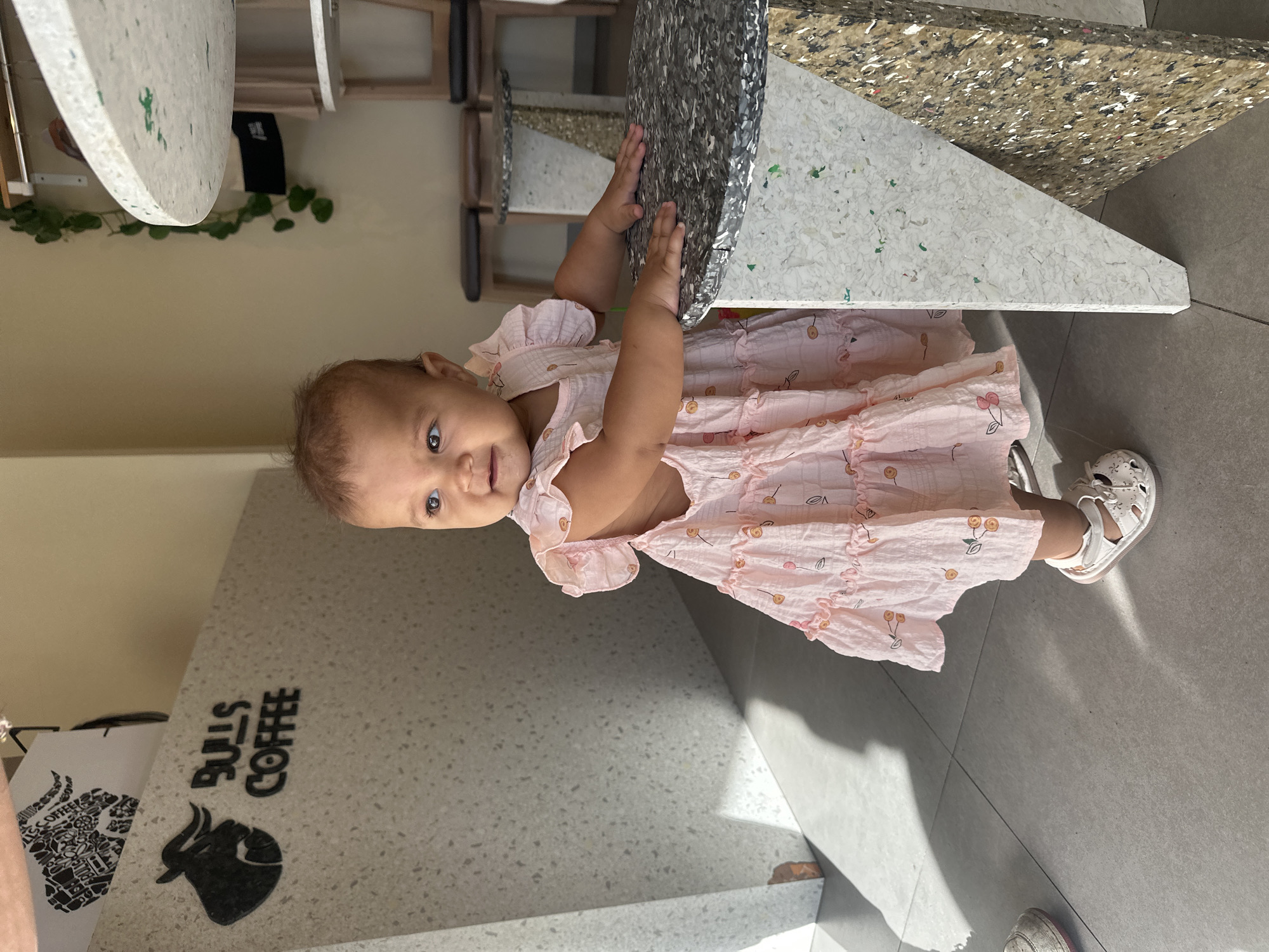 A young child is standing indoors, possibly in a cafe, as the logo "BULLS COFFEE" is visible on a countertop, indicating the business name. The child is wearing a pink dress with cherry patterns and white sandals. The background includes tables with unusual, speckled tops. The location might be North Kuta, Indonesia, based on provided information.