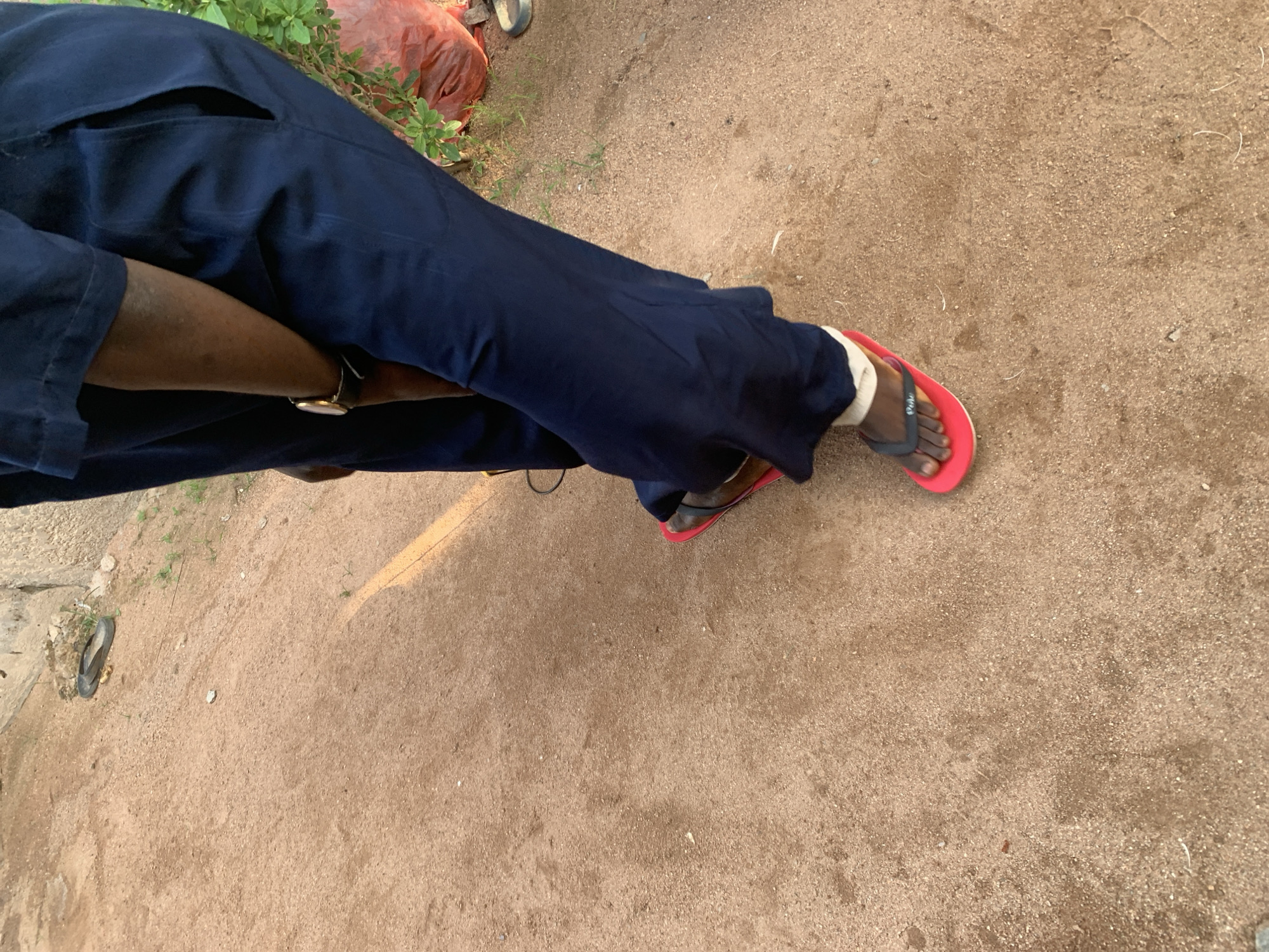 The image shows a close-up of a person's lower body, focusing on their legs and feet, which are wearing dark blue trousers and red flip-flops. The person is standing on a sandy, dirt path. A single black flip-flop is visible on the ground further up the path to the left. In the background, there is a rough, concrete structure and some green vegetation. A large, red plastic bag is also visible in the background to the right. The scene appears to be outdoors, possibly in a rural or semi-urban area, consistent with locations in Jalingo, Nigeria.