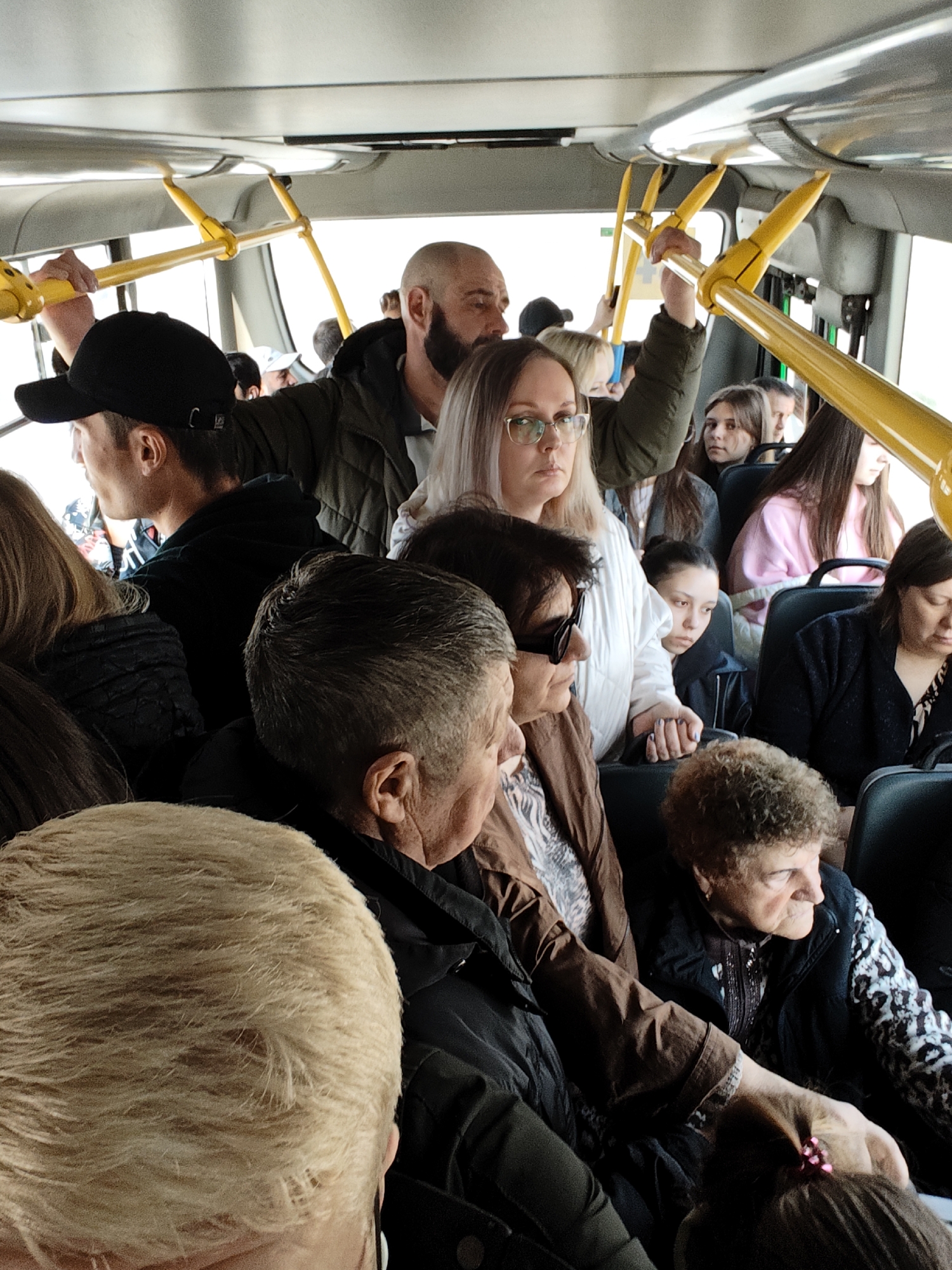 The image depicts a crowded public transportation bus, possibly in Yekaterinburg, Russia, given the context. The bus is filled with a diverse group of people of varying ages and appearances. Some individuals are seated, while others are standing and holding onto the yellow handrails for support. The lighting inside the bus appears natural, suggesting it's daytime. The environment seems confined due to the number of people present. No specific interactions or actions beyond traveling are prominently visible.