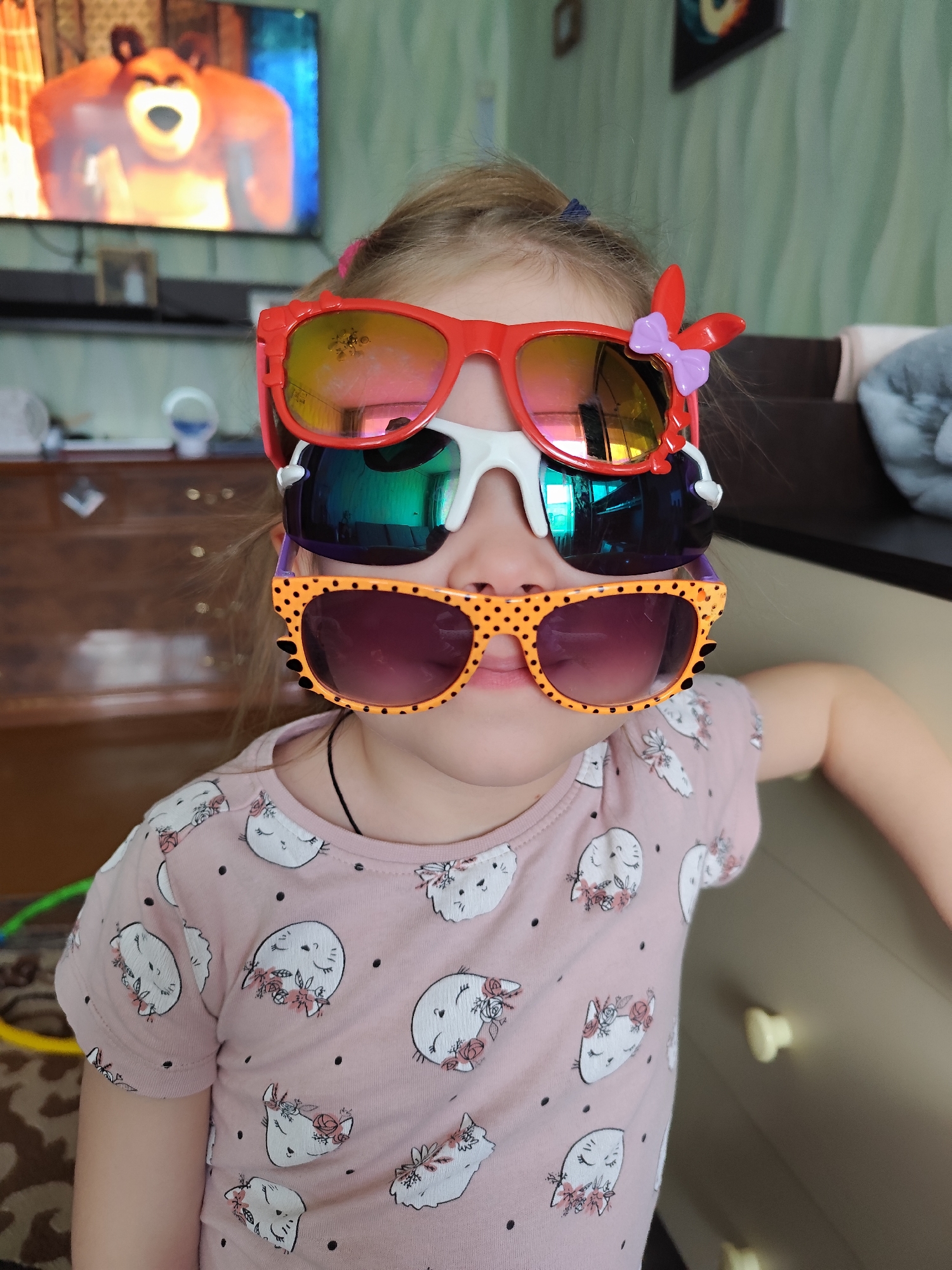 A young girl is indoors wearing three pairs of sunglasses stacked on her face. The top pair is red with a small purple bow on the side, the middle pair is white, and the bottom pair is orange with black polka dots. She is wearing a pink shirt with cartoon cat faces printed on it. Behind her is a wooden dresser, a television with a cartoon bear on the screen, and a brown sofa. The location is Фоки, Russia.