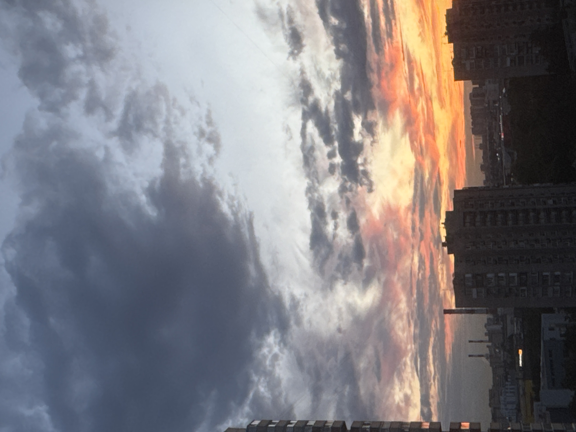The image depicts a dramatic sunset over an urban landscape in Saint Petersburg, Russia. The sky is filled with a swirling array of clouds, some dark and ominous, others illuminated by the setting sun with vibrant oranges, pinks, and yellows. This creates a stark contrast between the approaching storm and the warm hues of twilight.
Below the sky, the silhouette of a city is visible. Tall apartment buildings with numerous windows dominate the foreground and midground, their structures dark against the colorful sky. Some buildings appear to have lights on, suggesting evening activity within. In the distance, other structures, including what appear to be industrial chimneys or towers, are also visible, contributing to the dense urban feel of the scene. The overall impression is one of the raw power of nature meeting the built environment.
The time of day is clearly sunset, characterized by the low angle of the sun and the intense colors in the clouds. The weather appears to be dynamic, with evidence of both storm clouds and the colorful glow of a clear sunset. The location cues of apartment blocks and potentially industrial structures are characteristic of a large city. The dramatic and contrasting colors of the sky evoke a sense of awe and perhaps a touch of melancholy or anticipation, reflecting the transitional nature of the time of day and the weather. There is no visible text in the image.