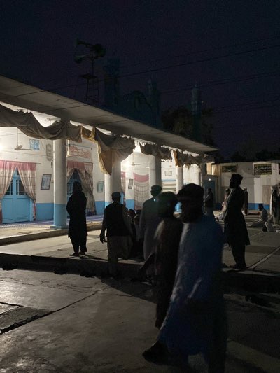 At dusk or night, a group of males are congregating on the grounds of a building in Kandahar, Afghanistan. The building has a light-colored facade with supporting columns and a roof extending out to provide a covered area. Some light is emitted from within the building, illuminating the area under the roof. Draped fabric hangs from the roof. Several of the males are standing near the edge of the covered area, while others are walking. The ground is paved and reflects the dim light. In the background, there are other buildings or structures, and the sky is a dark blue.