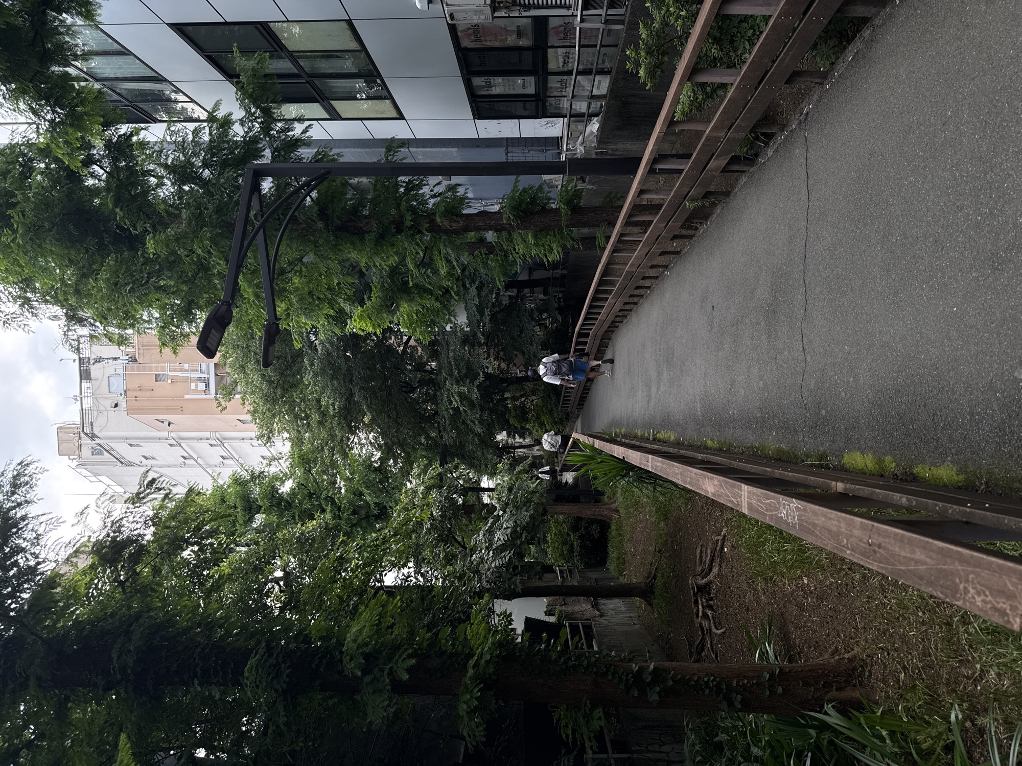 The image shows a paved pedestrian walkway in Shinjuku, Japan, bordered by a wooden railing on the right and a channel with vegetation and trees on the left. In the background, there are multi-story buildings and streetlights. One person, wearing a white shirt, blue shorts, and a backpack, is walking away from the camera down the walkway. Another person is visible further ahead, also walking away. The environment appears to be an urban park or a landscaped area within a city.