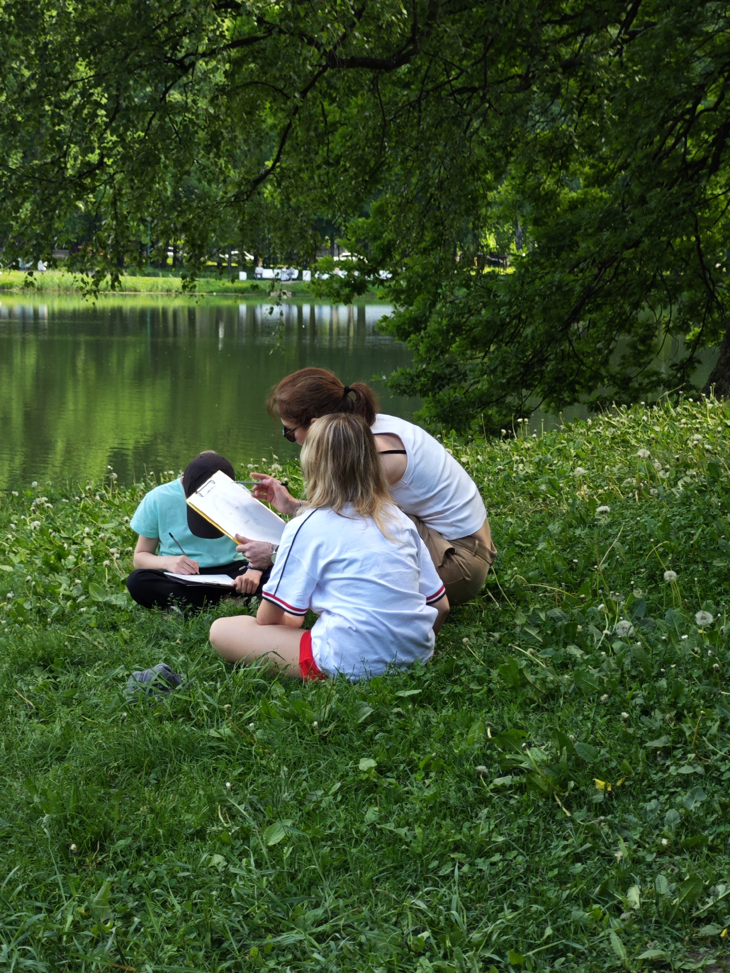 Three individuals are seated on a grassy area near a body of water, likely a lake or river, in Saint Petersburg, Russia. The setting is outdoors, with lush green grass and trees providing a natural backdrop. The person on the left, wearing a light blue t-shirt and black pants, is looking down at a clipboard and appears to be writing or drawing with a pen. The person in the middle, a woman with brown hair, is leaning forward, holding a clipboard with a white paper and pointing at it with her left index finger, possibly explaining something. She is wearing a tan skirt and a white top. The third person, on the right, is a young woman with blonde hair, wearing a white t-shirt and red shorts. She is facing away from the camera, towards the other two individuals. The water behind them is calm and reflects the green foliage. Dandelions are scattered throughout the grass. The overall impression is that of an outdoor lesson or study session.