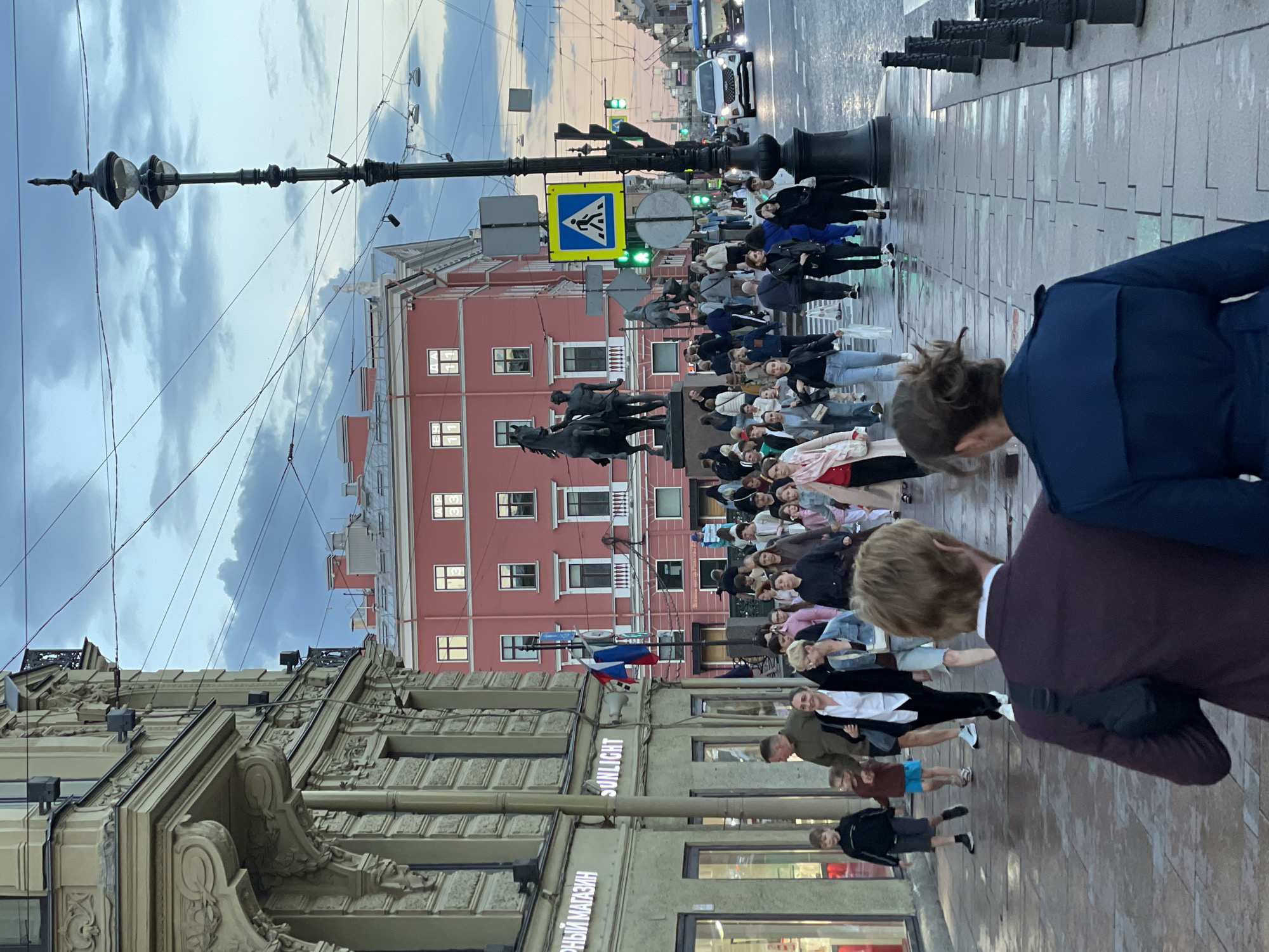 The image captures a street scene in Saint Petersburg, Russia, likely during the evening or late afternoon due to the subdued lighting and overcast sky. A wide sidewalk is filled with a diverse crowd of pedestrians walking in various directions. Several people are in the foreground, facing away from the camera, including a man in a burgundy jacket and a woman in a blue coat. Behind them, a dense flow of people moves along the sidewalk.
On the left side of the frame, a historic building with ornate architectural details and large display windows is visible. Signs indicate a store with Cyrillic text and "SUNLIGHT." To the right of the sidewalk, a busy street features a white car and a blue bus. Traffic lights are visible, with one showing green. A distinctive black lamppost with multiple globes stands prominently in the middle ground.
In the background, a pink building with many windows stands out. In front of this building, a large equestrian statue is mounted on a pedestal. A blue, white, and red flag is affixed to a pole near the statue. The ground appears wet, suggesting recent rain. The overall atmosphere is one of urban activity and movement within a historic city setting.