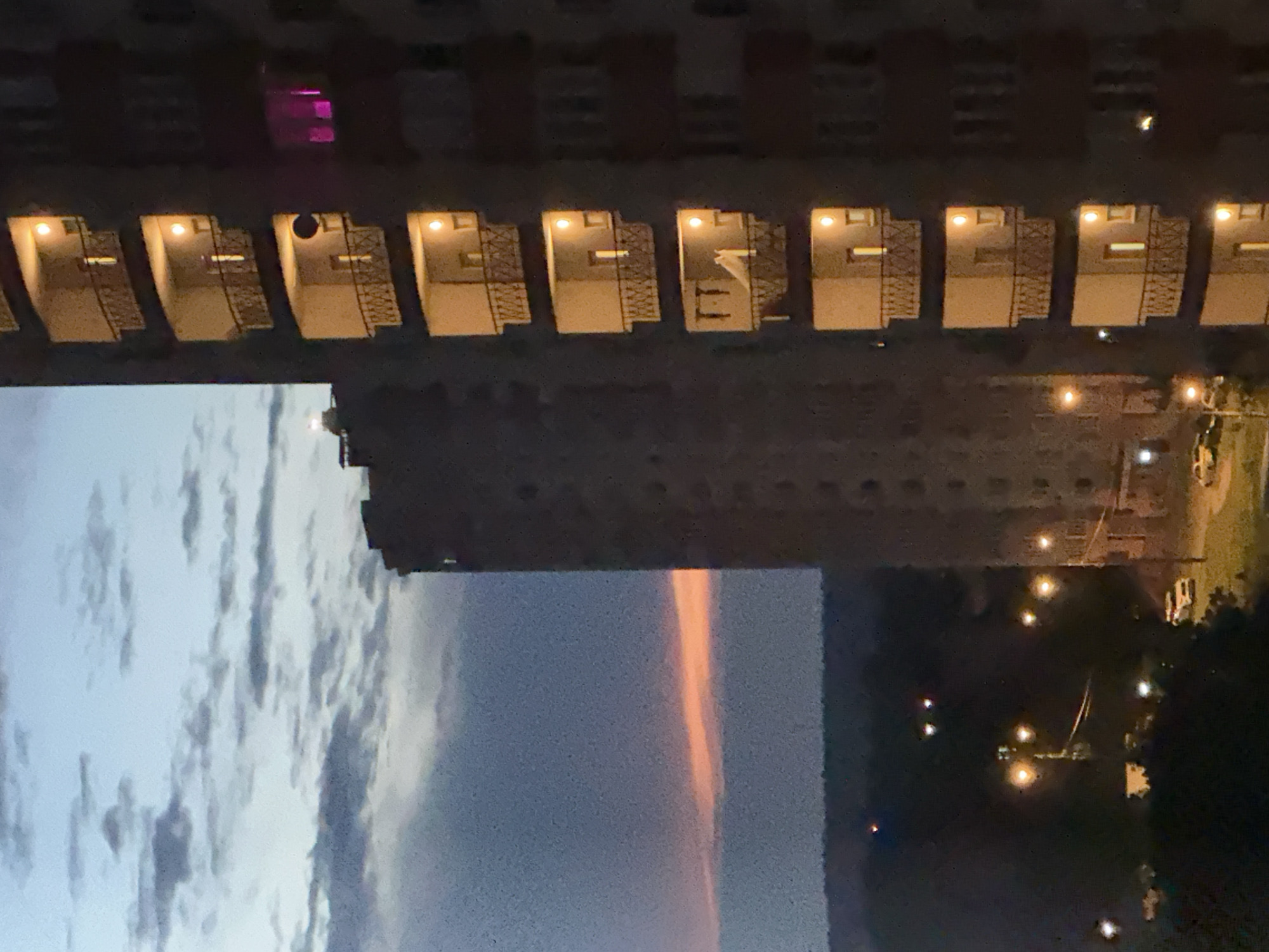 This is a medium-shot, eye-level, vertical photograph of a tall apartment building at dusk. The sky is a gradient of blue and gray with a thin band of orange light on the horizon. Several balconies are visible on the right side of the building, each lit from within. One balcony on the upper right has a pinkish glow. The lower part of the image shows a street with a few cars and streetlights. A second, slightly shorter building is visible to the left of the main apartment building. The scene appears to be in a residential area of a city.