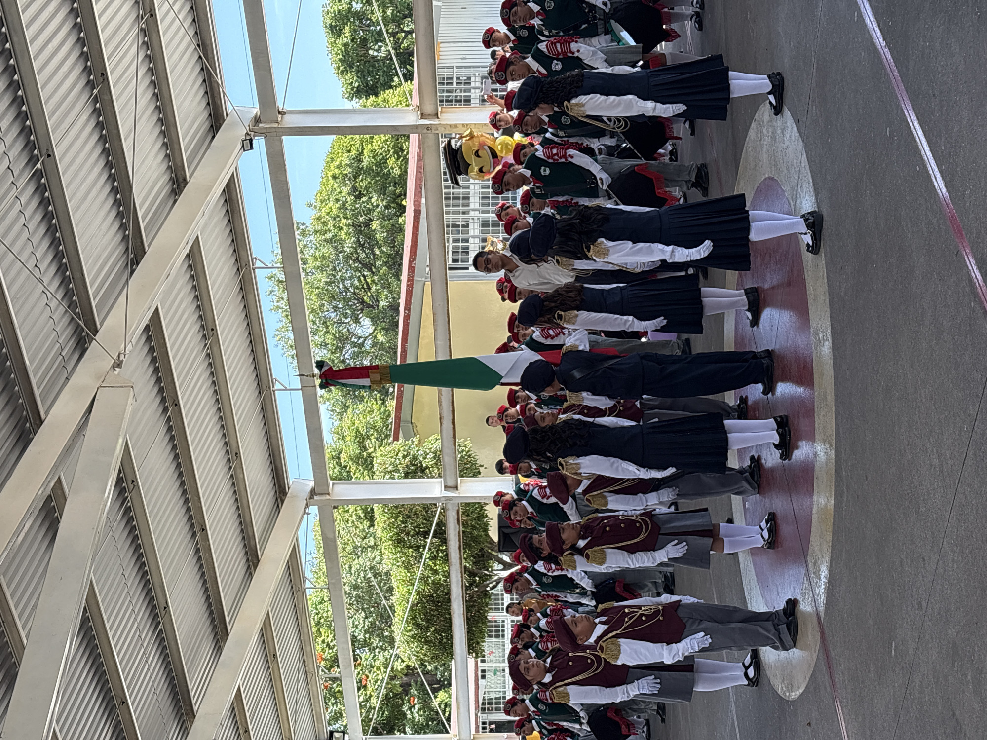This is a full-shot, eye-level photograph of a school marching band ceremony taking place outdoors in Tejalpa, Mexico. The scene is covered by a metal roof structure supported by white pillars. In the foreground, a group of students in uniform, mostly wearing burgundy berets and vests over white shirts and dark skirts or pants, stand at attention. Some are holding drums. A Mexican flag on a pole is visible to the right of the frame. In the background, more students are gathered, along with a few adults. One adult, wearing glasses and a light-colored shirt, stands near the flag. To the right, a large, yellow balloon resembling a graduation cap and a smiling emoji character floats in the air. The ground is a paved surface with circular markings. Lush green trees and buildings are visible beyond the covered area. The overall atmosphere appears to be one of formality and pride, likely for a school event.
