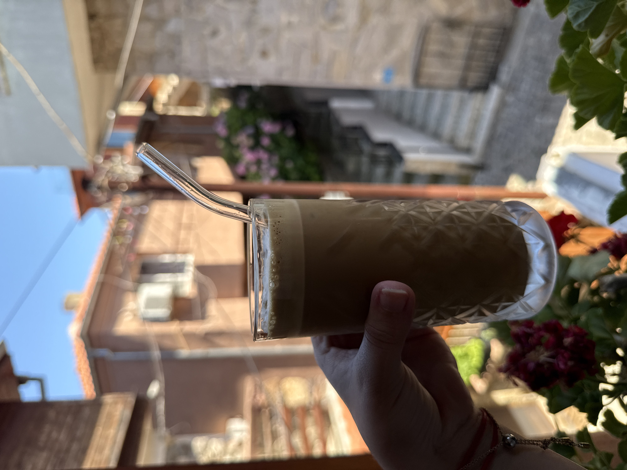 A person's hand holds a clear glass containing a cold coffee drink with foam and ice. A glass straw is inserted into the drink. The hand has a red bracelet on the wrist. The background is out of focus but shows a sunny day in a European town, possibly Sozopol, Bulgaria. Buildings with terracotta roofs and balconies are visible, along with steps leading up a street. Green foliage and flowers are present on the right side of the image. The overall impression is one of a refreshing drink enjoyed outdoors on a pleasant day.