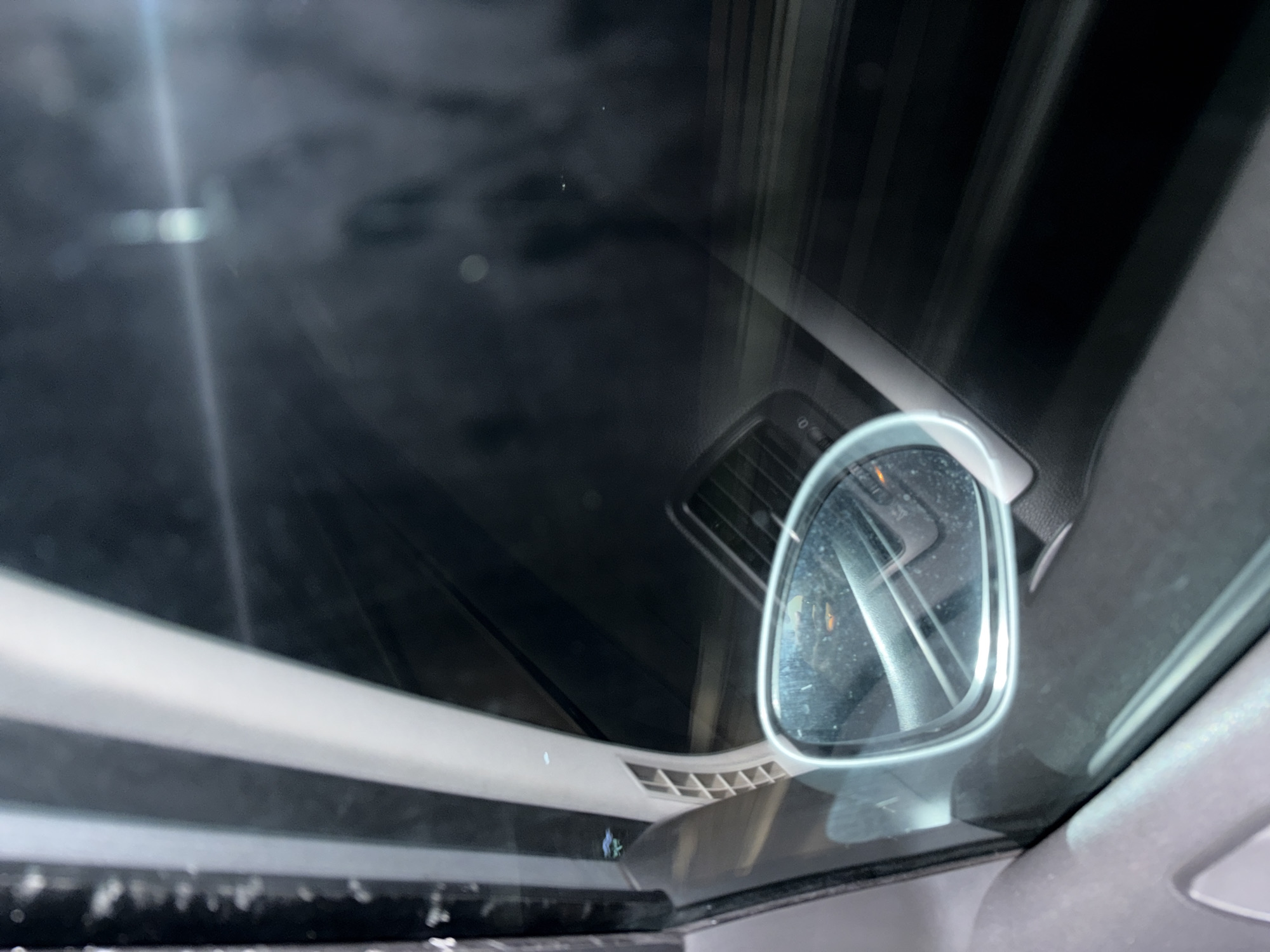 A car's interior is viewed from the driver's seat, focusing on the rearview mirror. The mirror reflects a blurred image of the car's interior. The car's roof and frame of the sunroof are visible. Through the windshield, the sky and blurred shapes are visible. The location is unclear and cannot be determined based on the image. There are no people or actions visible.