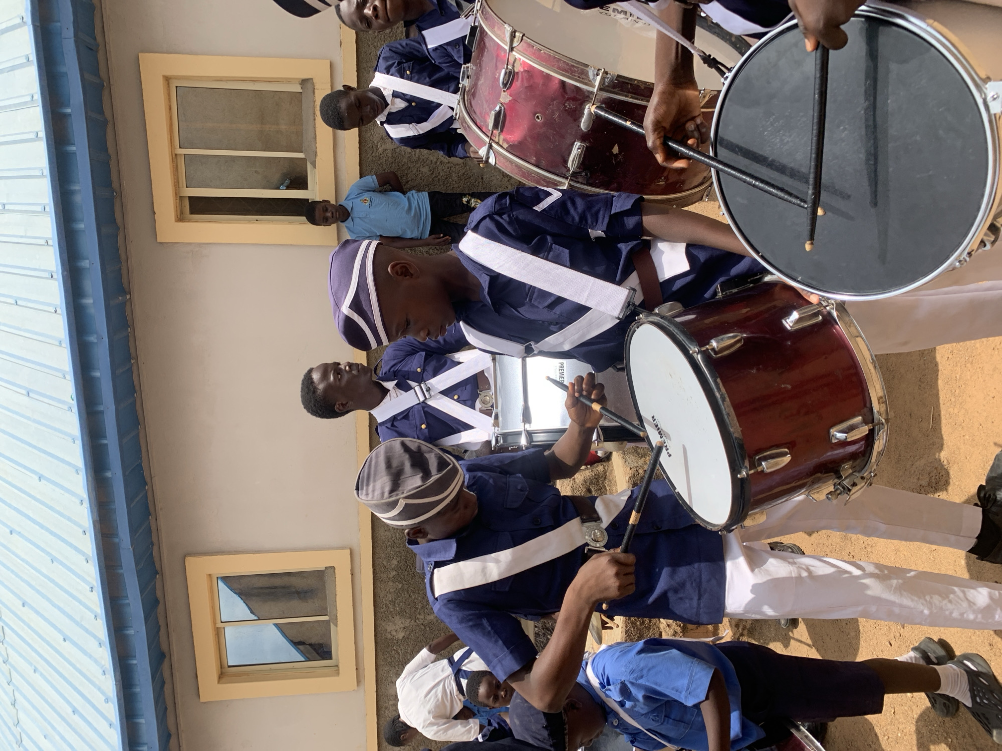 A group of young men dressed in matching navy blue uniforms with white diagonal straps and white trousers are playing drums outdoors. They are standing in formation, with drums of varying sizes, including bass drums and snare drums. Most of the individuals have matching navy and white striped caps. They are holding drumsticks and appearing to be actively performing. The environment is outdoors, in front of a light-colored building with framed windows. In the background, there is a boy in a blue shirt who may be watching. The location appears to be in Jos, Nigeria.