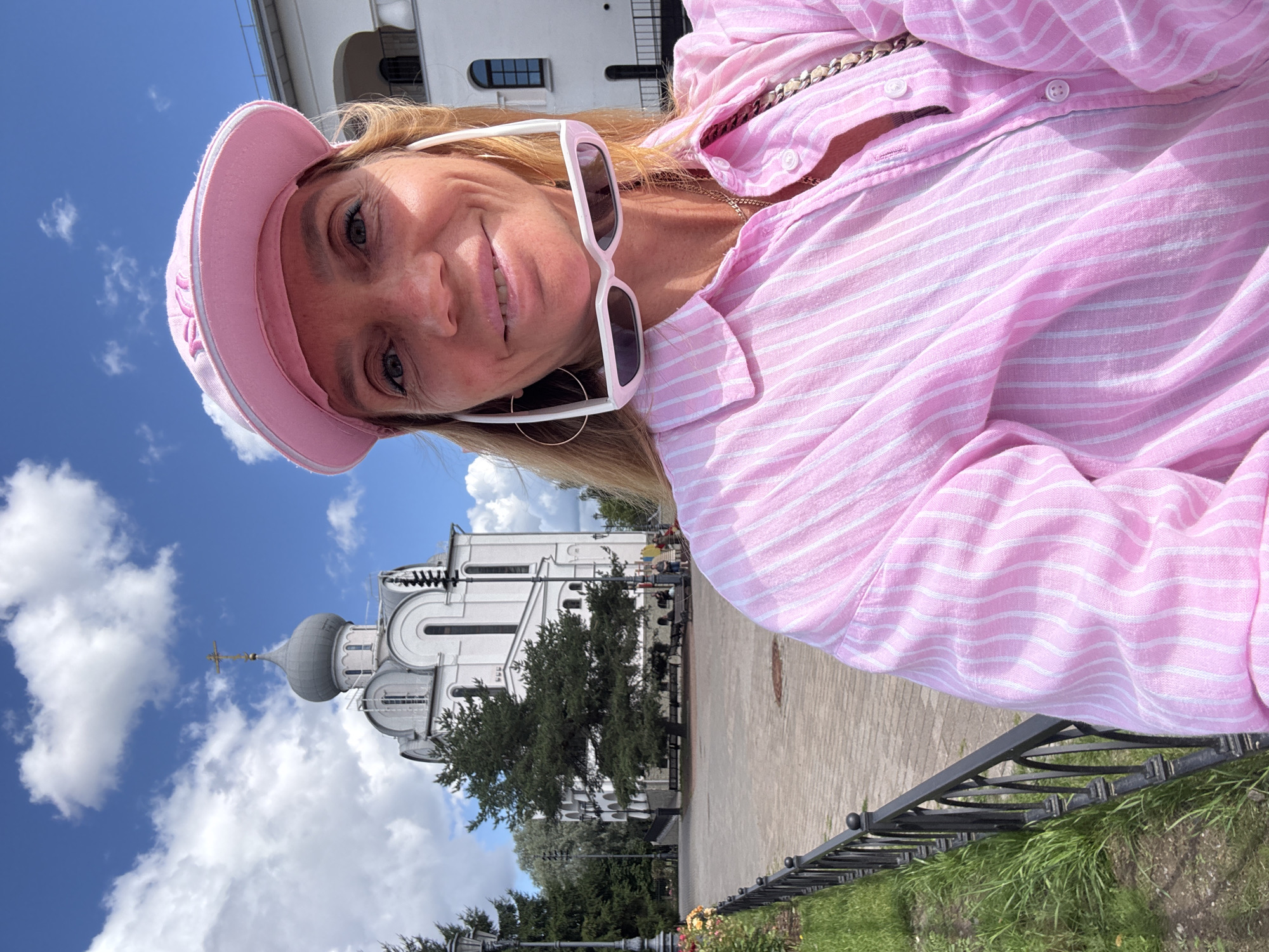 An outdoor selfie features a woman with blonde hair and sunglasses perched on her head. She is wearing a pink striped button-down shirt and a pink baseball cap. Behind her, a white church with a grey dome and a cross on top is visible, along with green trees and a blue sky with white clouds. The church is identified as the Church of the Resurrection of Christ in St. Petersburg, Russia.