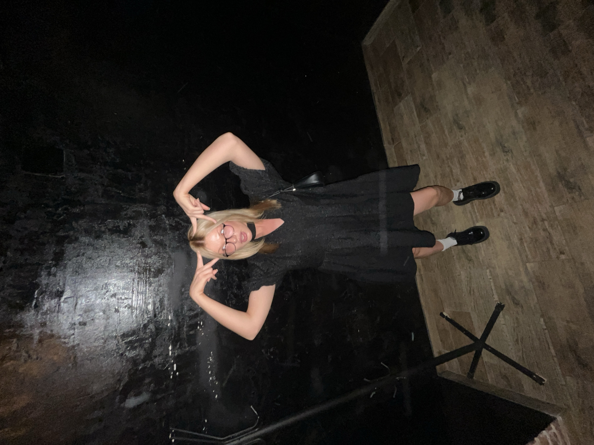 A young woman with blond hair and glasses is posing indoors. She is wearing a black dress and black shoes with white socks. She is making a hand gesture near her face. The setting appears to be a room with a mix of flooring: dark, possibly polished flooring on one side and lighter, wooden-looking flooring on the other. The background includes what looks like a metal structure. There is no information in the image to determine this is in the city of Chaikovsky, Russia.