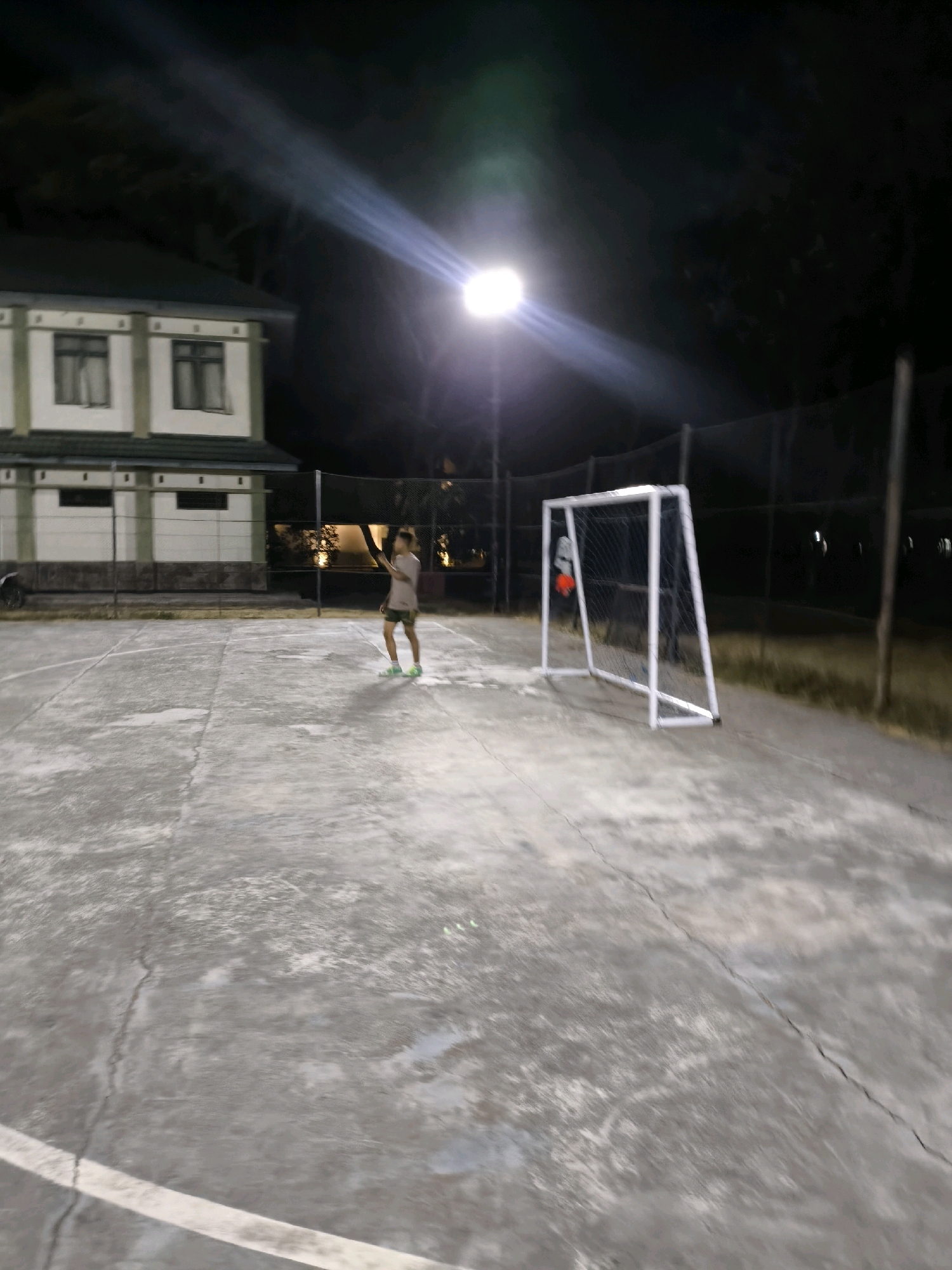 The image depicts an outdoor basketball court at night, illuminated by a bright overhead floodlight. The court surface is concrete, showing visible cracks and white boundary lines. In the background, a two-story building with green trim and white walls is visible. A person, appearing to be a young male wearing a light-colored t-shirt and dark shorts with green accents, is standing on the court, holding a racket and facing away from the camera. To the right of the person, a white soccer goal with a net is set up on the court. The scene is enclosed by a chain-link fence, and beyond that, trees and darkness suggest a natural environment surrounding the court.