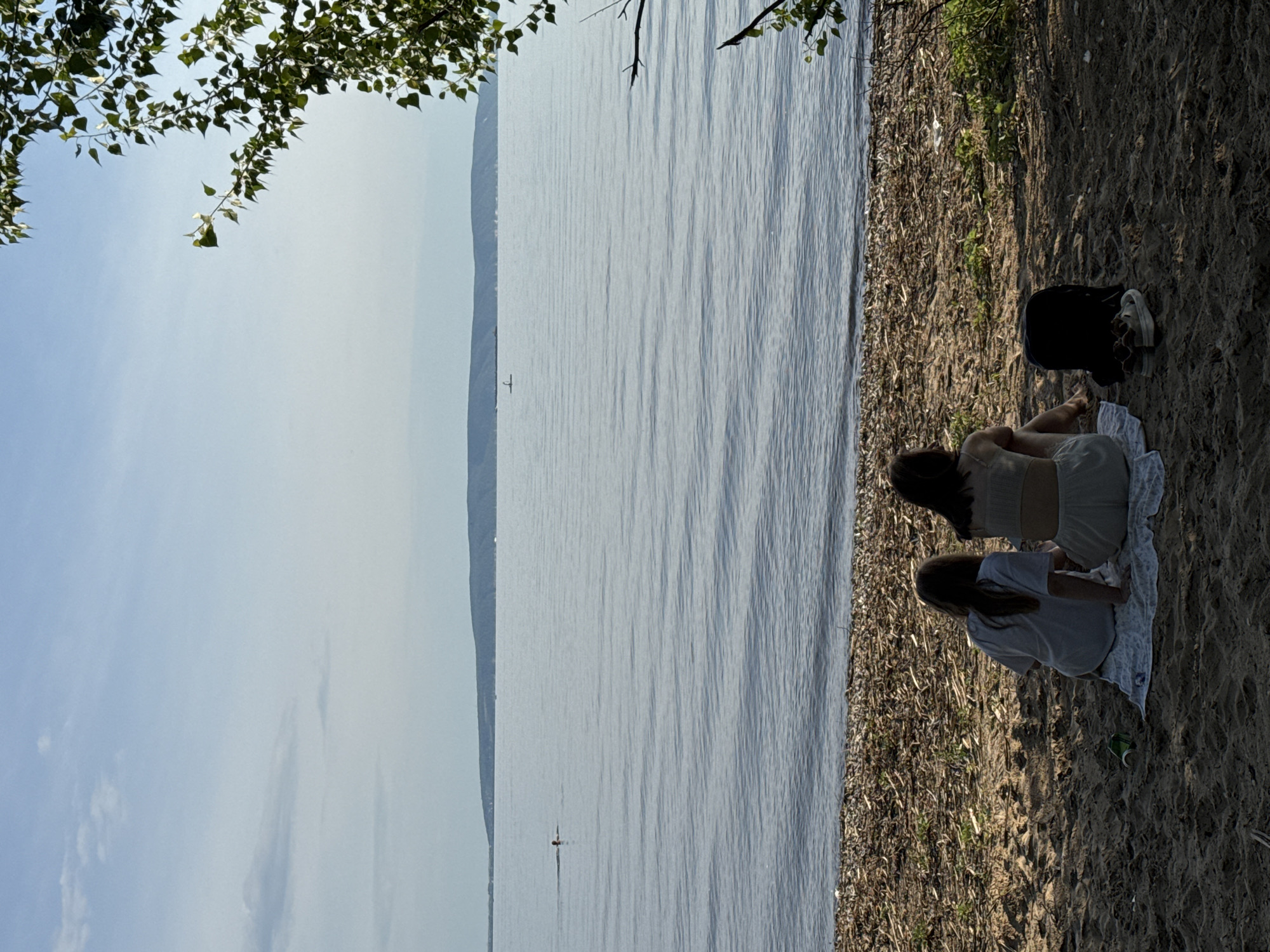 The image shows two young women sitting on a sandy shore next to a large body of water. One woman is wearing a light-colored top and shorts, and the other is wearing a light-colored t-shirt. They are both sitting on a white blanket. A dark bag and a pair of shoes are on the sand behind them.
The scene appears to be a relaxed, outdoor setting. The water is calm with gentle ripples, and a distant shore is visible across the water. A single small boat with a person in it is also visible on the water. Tree branches with green leaves hang from the top left of the frame, suggesting an outdoor, possibly natural environment. The lighting suggests it is daytime, likely late afternoon or early evening due to the shadows on the sand. The overall mood seems peaceful and serene. There is no visible text in the image.