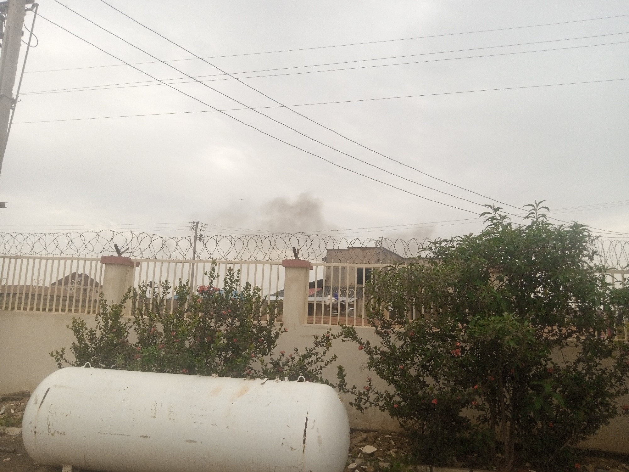 A large white propane tank lies horizontally on the ground in the foreground of the image. Behind the tank, a light-colored fence topped with barbed wire stretches across the frame. Beyond the fence, several buildings are visible, with a plume of dark smoke rising into the overcast sky from behind one of them. To the right of the propane tank and in front of the fence, there are two large, leafy green bushes with small pink flowers. Power lines are visible in the upper portion of the image, stretching across the sky. The scene appears to be in an outdoor urban setting, likely in the city of Jos, Nigeria, given the presence of the propane tank and architectural styles of the buildings, coupled with the commonality of power lines in urban environments. The smoke suggests a potential fire or industrial activity.