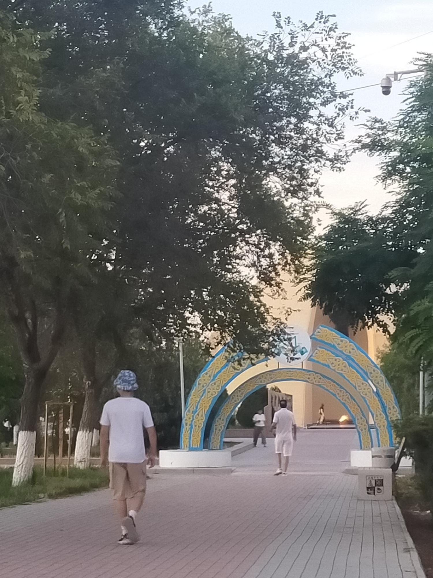 The image shows a person walking away from the camera on a paved walkway in an outdoor urban setting. The person is wearing a white t-shirt, khaki shorts, and a blue and white bucket hat. To the right of the walkway, there are trees and foliage. In the background, there is a large, ornate archway with blue and yellow decorative elements. The archway appears to be made of metal and has some symbols or text on it. Behind the archway, a building is visible. Another person is walking away from the camera in the distance, under the archway, and a third person is walking towards the archway on the other side. A surveillance camera is visible mounted on a pole among the trees to the right of the image. The sky is visible above the trees and appears to be cloudy or overcast.