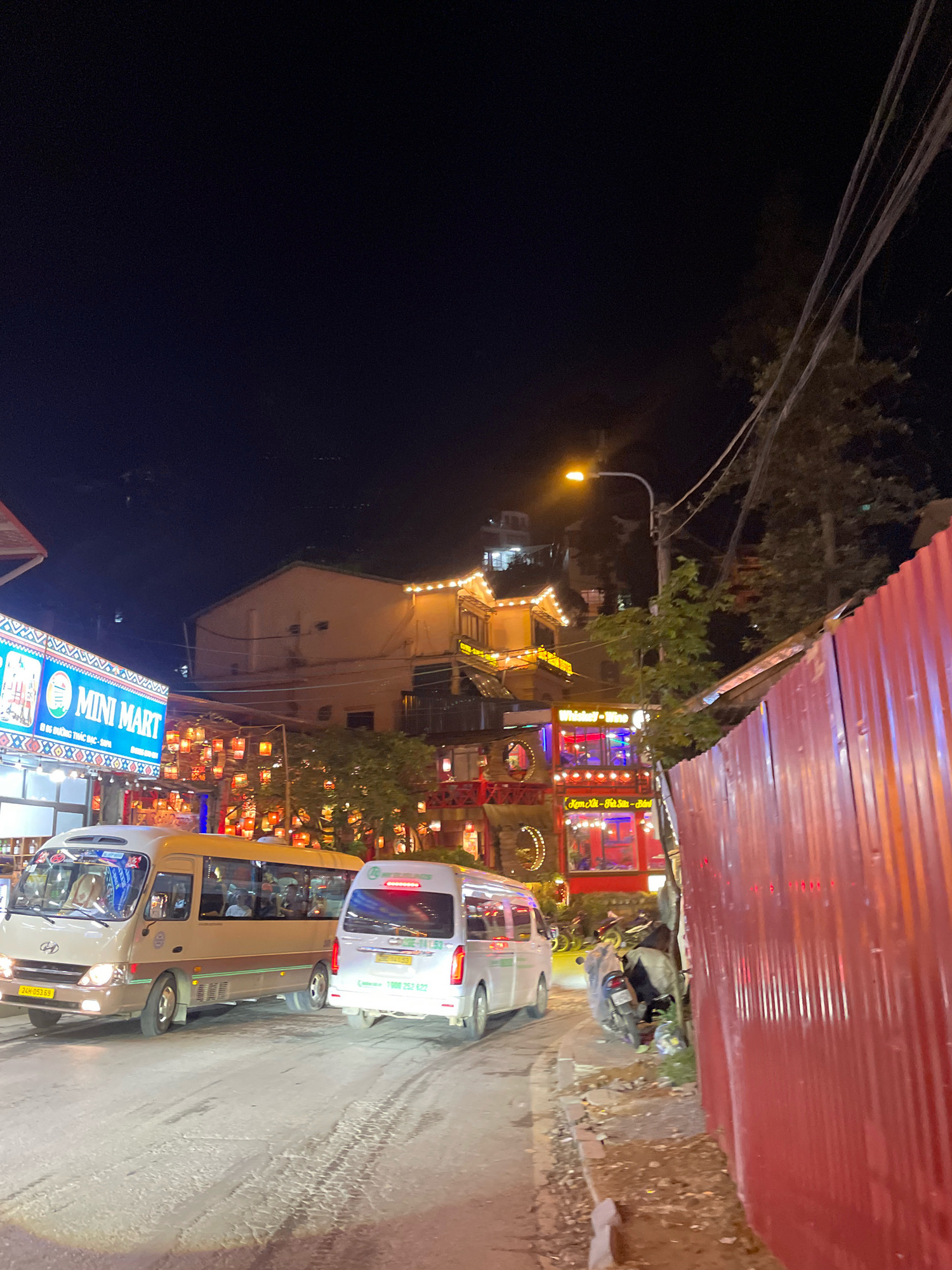 The image shows a street scene at night in Sa Pa Ward, Vietnam. Two minivans are driving on a paved road. The minivan on the left is beige and has the license plate "24H 051.65". The minivan on the right is white and has a red taillight illuminated; its license plate is partially visible and appears to be "7006-011.63". To the left of the road, there is a building with a sign that reads "MINI MART". Across the street, a multi-story building is illuminated with festive string lights, and numerous red lanterns are hanging from trees and structures, creating a vibrant atmosphere. A motorcycle is parked near the right side of the road, next to a corrugated red metal fence. The sky is dark, indicating it is nighttime.