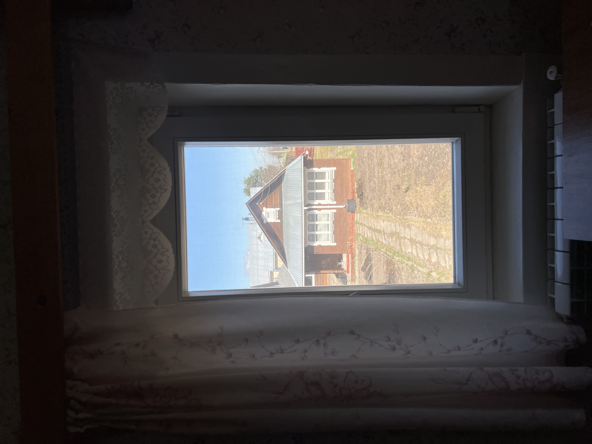 The image depicts a view from a window of a house in Чернышово, Russia. Outside the window, another house with a grey roof and brown siding is visible, as well as bare patches of ground. A faint walkway leads to the house. The sky is light blue. Inside, the window is draped with patterned curtains, and a radiator can be seen on the right. The overall scene appears to be taken during the day, likely early spring or late fall.