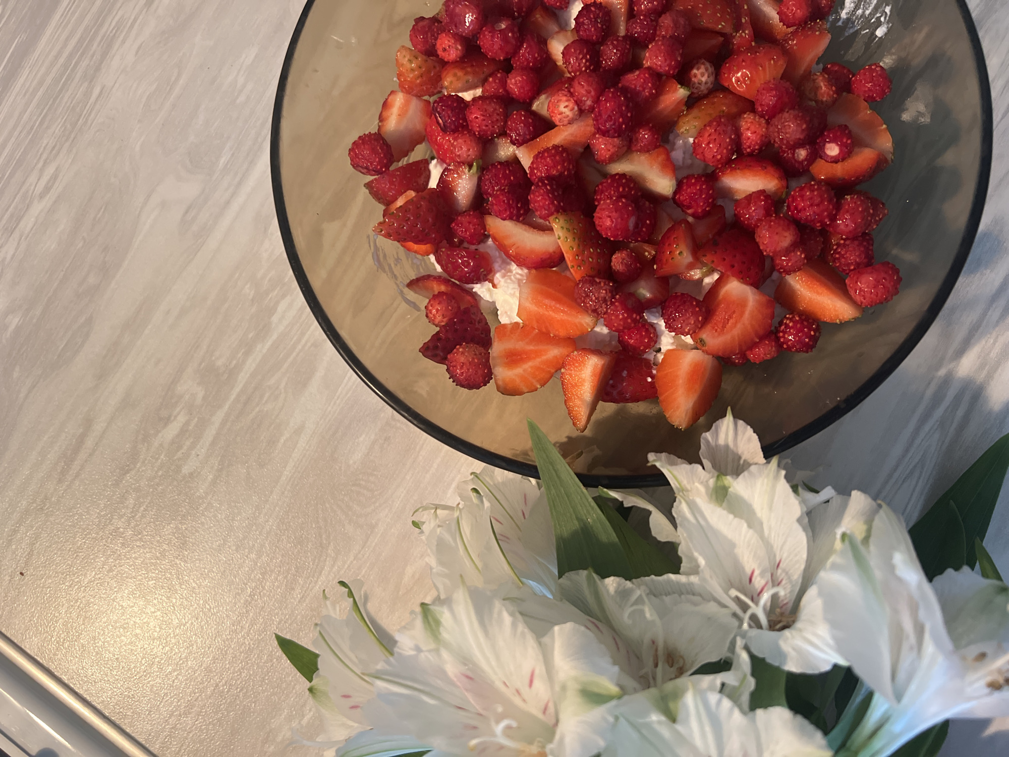 A close-up, overhead shot shows a bowl of strawberries and wild strawberries mixed with cream, placed on a light-colored, wood-patterned surface. To the left, a bouquet of white alstroemeria flowers with pink and green accents is visible, some of their leaves extending towards the bowl. The lighting suggests an indoor setting, possibly a kitchen or dining area. There are no people or specific landmarks indicating a location in Chaikovsky, Russia.