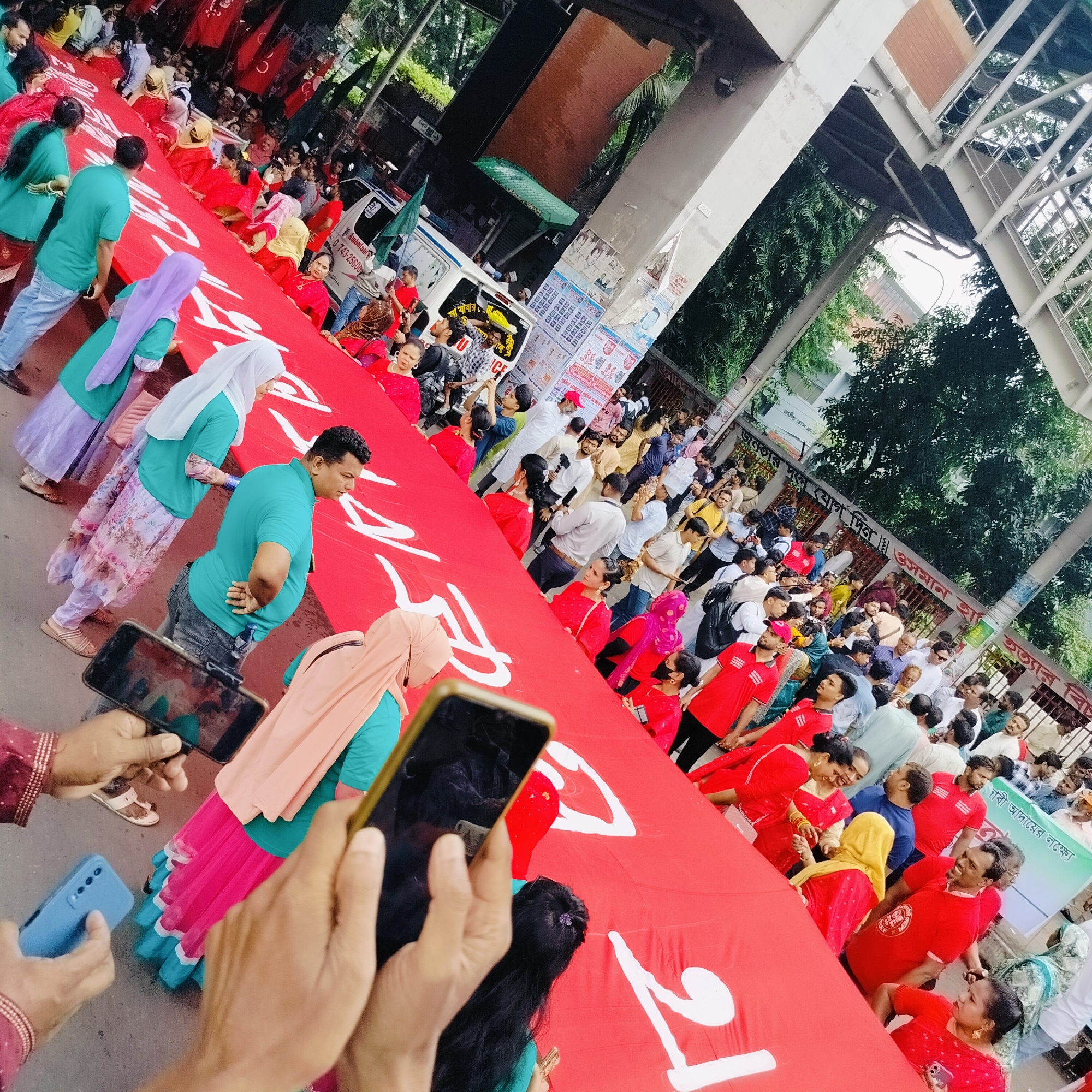 A wide-angle, high-angle shot depicts a crowd of people in what appears to be a protest or demonstration in Dhaka, Bangladesh. A large red banner with white lettering is being held by numerous participants. Many individuals are wearing red shirts, while others are dressed in varied colors, including shades of blue, green, and white.
Several people are holding up their mobile phones, presumably to take photos or videos of the event. An ambulance is visible in the background, parked near the crowd. Above the people, an elevated pedestrian walkway or overpass spans the scene. The surroundings include buildings, foliage, and posters affixed to structures. The text includes Bangla words and numbers, further indicating the location.
