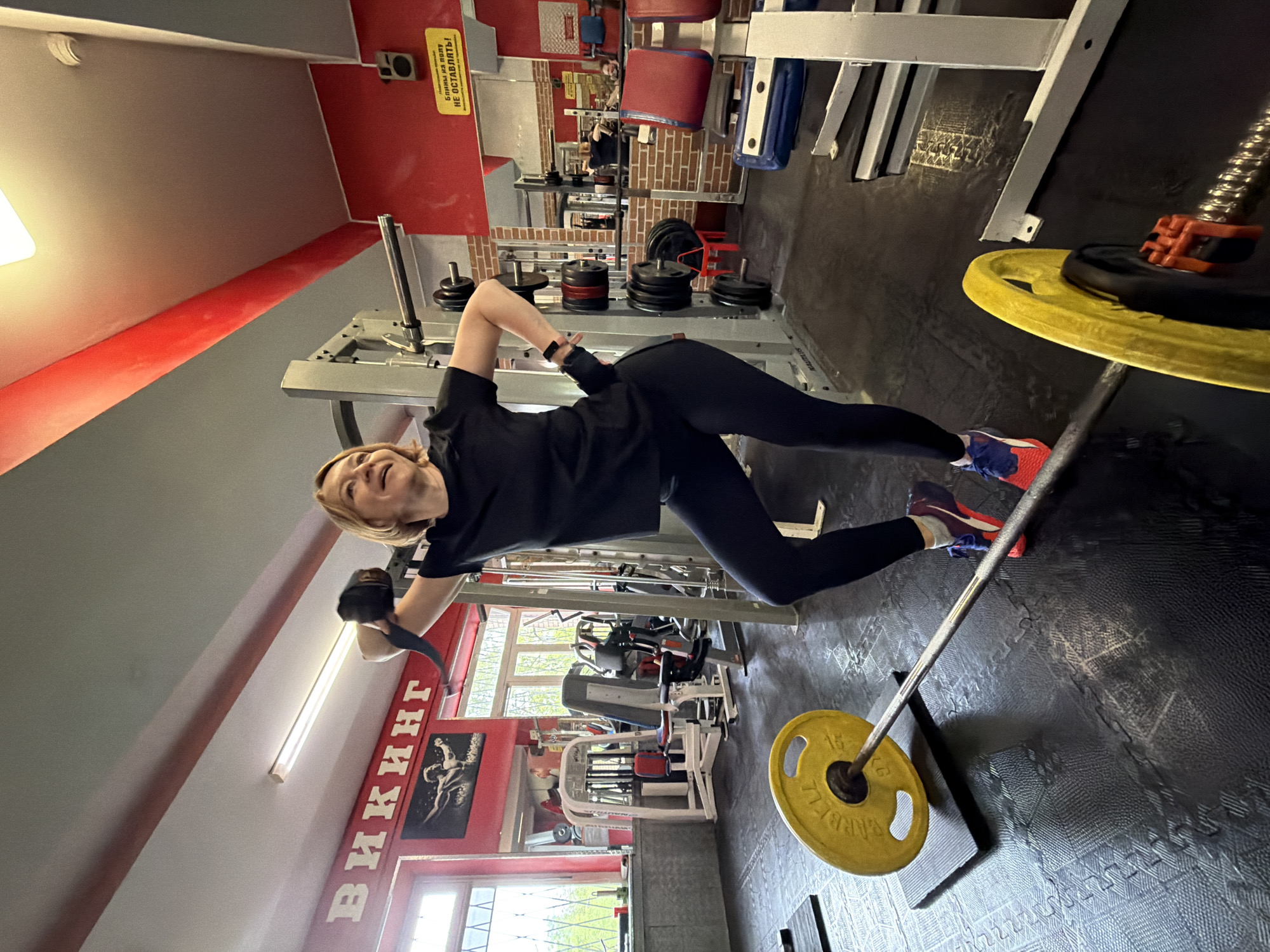 A woman is performing a core workout at the "VIKING" gym, located in Saint Petersburg, Russia. She is suspended from a pull-up bar using straps, holding a plank position. She wears a black shirt, black leggings, and colorful sneakers. The gym features various weightlifting machines and equipment, including a squat rack, a cable machine, and a barbell with yellow weight plates. The gym has red and gray walls with motivational posters. The floor is covered in black rubber mats.