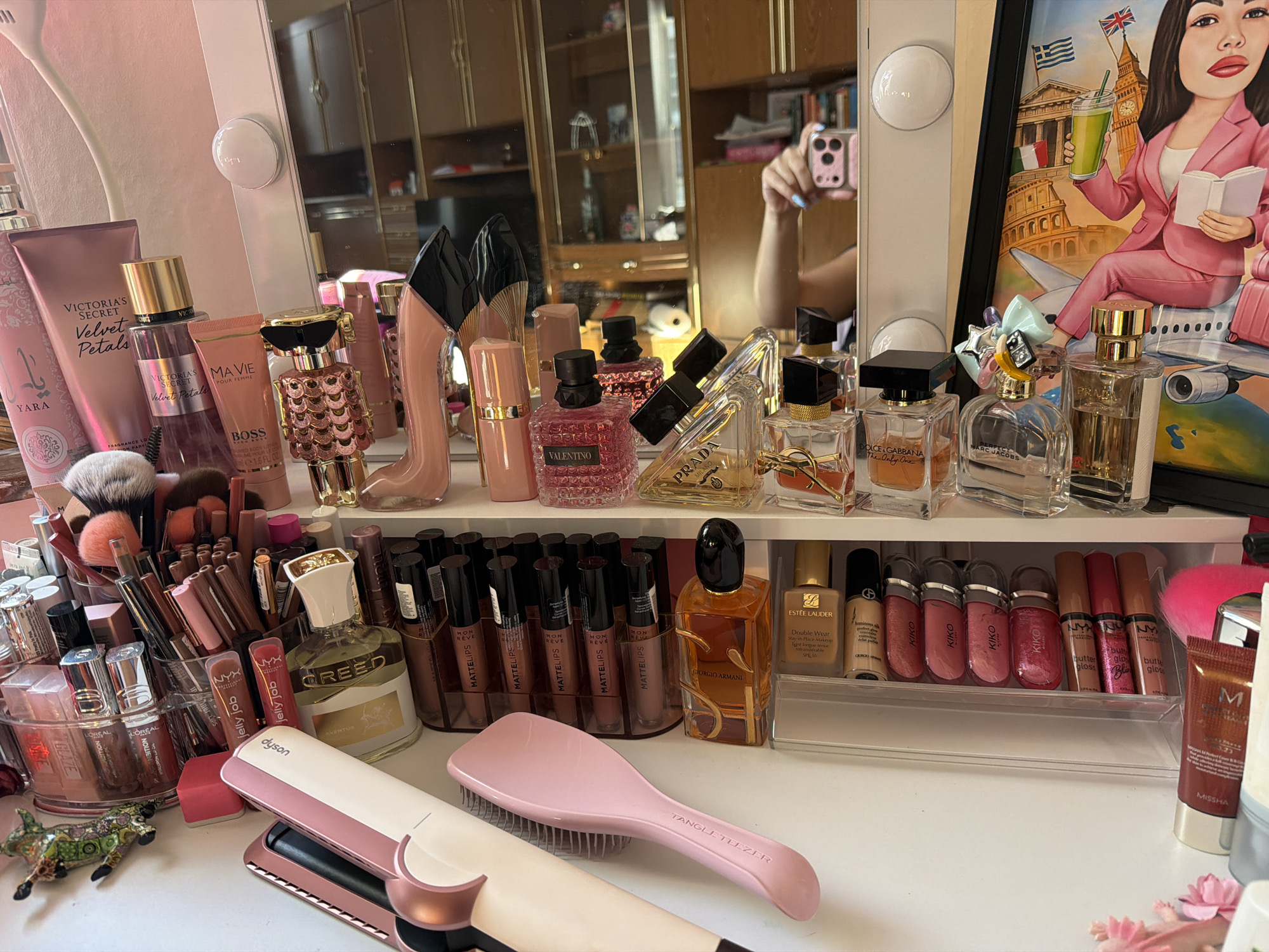 The image shows a vanity filled with a collection of beauty products, including perfumes, makeup, and hair styling tools. A person is visible in a mirror in the background, holding up a smartphone to take a selfie. The overall setting appears to be a personal dressing area, likely a bedroom or bathroom, with various shelves and drawers visible. The lighting suggests it is daytime, with artificial lights integrated into the vanity mirror as well. There is no indication of the city of Schwelm, Germany, or any specific location within it.