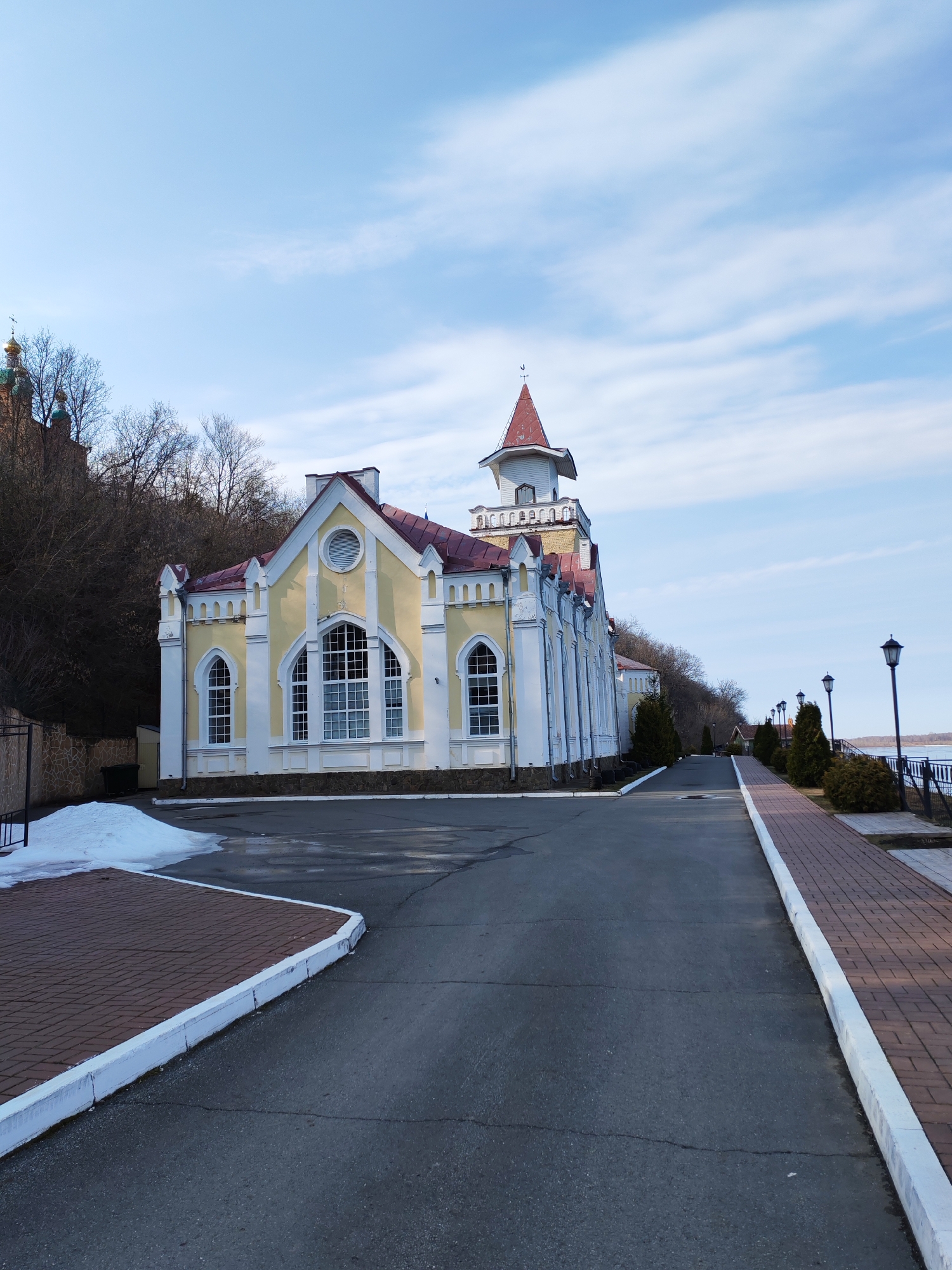 The image captures a bright, sunny day in Sarapul, Russia. The focal point is a yellow and white building with a red roof and a small tower. The building is situated next to a street and a raised walkway made of brown bricks. A small patch of melting snow is visible near the building. In the background, trees cover a hill, with glimpses of a building with green domes and a cross. The sky is blue with scattered clouds. The street leads towards a row of lampposts and a body of water.