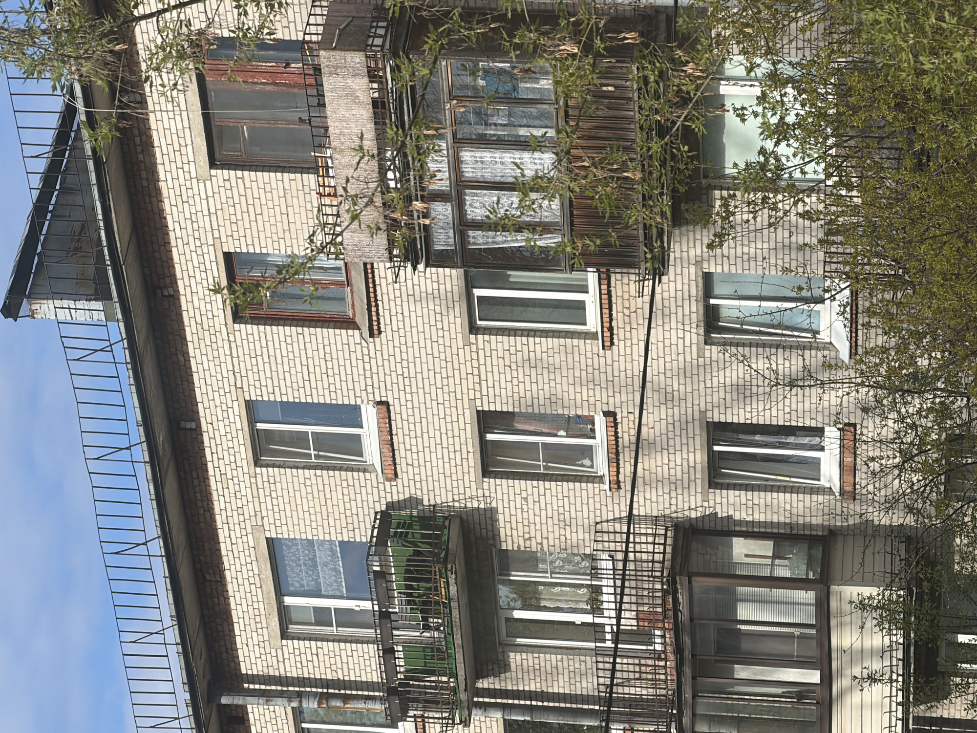 The image shows the facade of a multi-story brick building with multiple windows and balconies. Some windows have curtains visible. A fire escape ladder is attached to the side of the building. The building appears to be in a state of disrepair. The presence of trees suggests an urban or suburban location. The location is specified as Tokkari, Russia. No people are visible in the image.