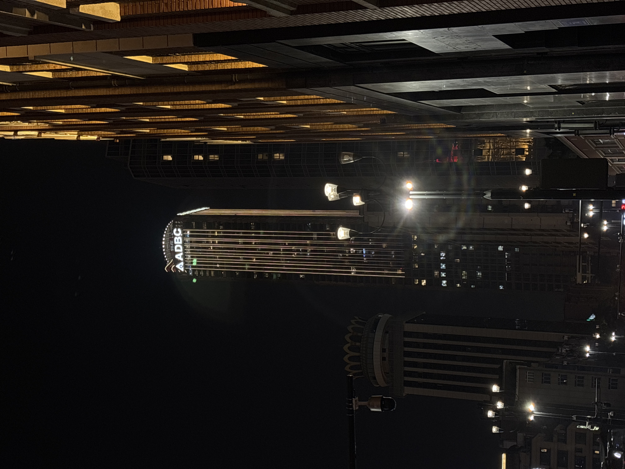 The image shows a night scene in Yuzhong District, Chongqing, China. The prominent building is the AADBC building, identifiable by its sign. Streetlights are visible, casting light onto the surrounding buildings. The buildings exhibit lit windows. The scene is dark, indicative of nighttime.