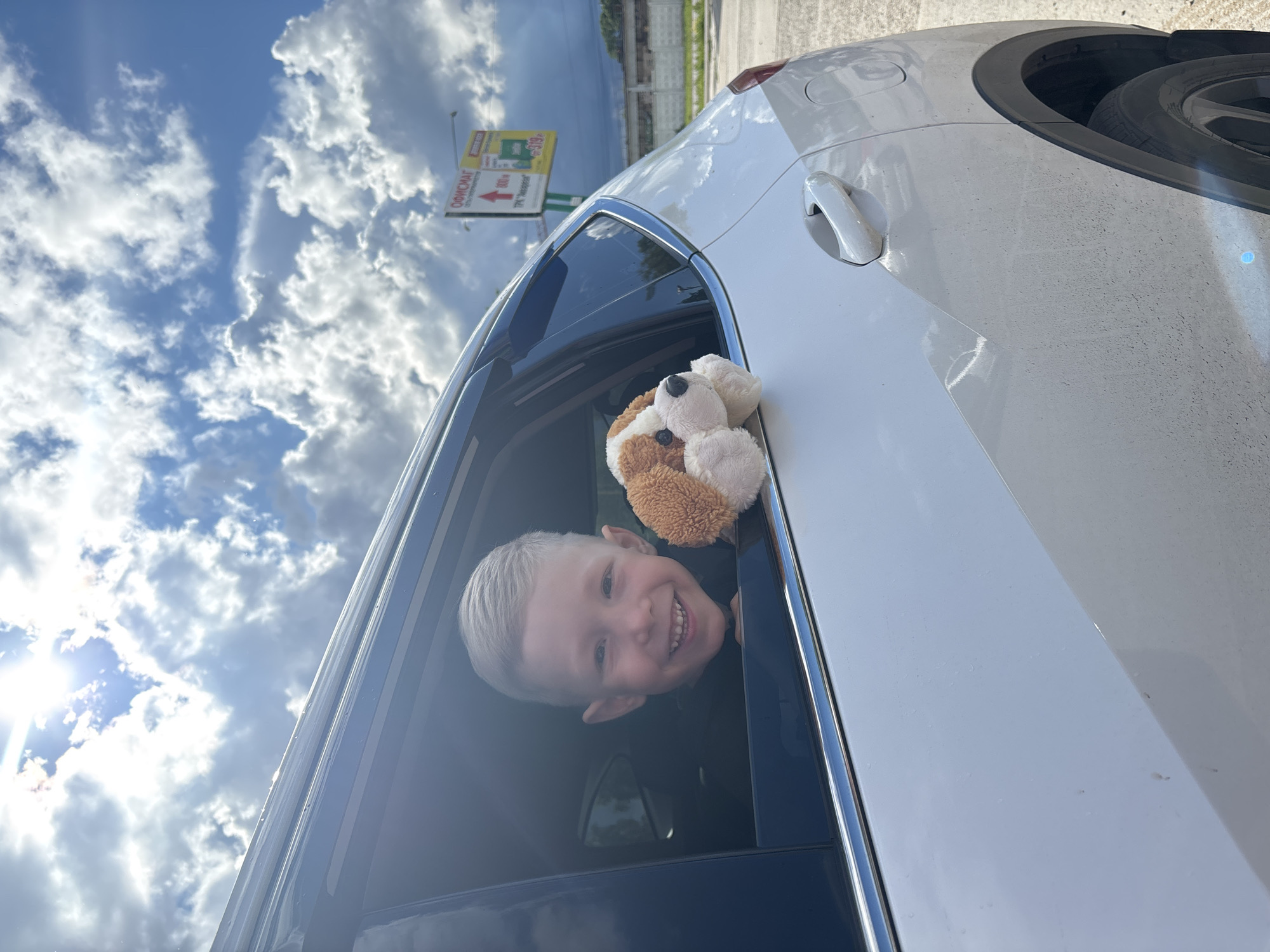 A cheerful young boy with blonde hair is smiling broadly and peeking out of the open window of a white car. Next to him, a brown and white stuffed dog or puppy toy is also visible, propped in the car window. The boy's happy expression suggests a playful or joyful moment.
The scene takes place outdoors on a bright, sunny day with a blue sky filled with white clouds. Sunlight is strong, causing a lens flare effect on the left side of the image. The car appears to be a modern, clean white vehicle, possibly an SUV or hatchback, with chrome trim around the window.
In the background, an advertising billboard is visible, featuring Cyrillic text. Notable text includes "ОФИСМАГ" (OFISMAG) and "ТЦ 'Якорь'" (TC 'Yakоr', meaning Shopping Center 'Anchor'), along with an arrow pointing left and numbers like "800" and "519". These details, combined with the provided context, confirm the location as Chaikovsky, Russia.