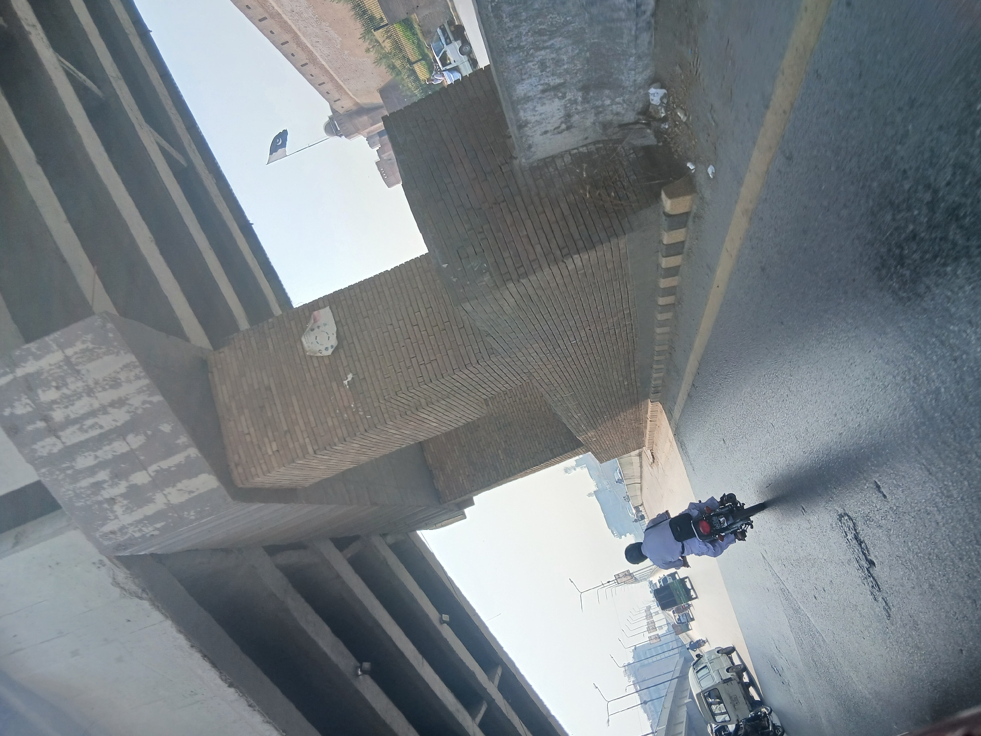 A street-level, wide-angle, slightly tilted shot captures a scene in Peshawar City Tehsil, Pakistan. The sky is bright and hazy, suggesting it is daytime. In the foreground, a person on a motorcycle wearing a white shalwar kameez and a black helmet is moving away from the viewer on a paved road. The motorcycle is black with a red taillight. Behind the motorcyclist, several vehicles are visible on the road, including a white van, a green truck, and other cars and motorcycles.
Dominating the upper portion of the frame is a concrete overpass with multiple beams, casting shadows on the road below. To the right of the overpass support structure, a large, weathered brick pillar is visible, part of the overpass's infrastructure.
Further in the background, a portion of a historic fort-like structure with crenellated walls and domed turrets is seen. A flag, appearing to be the flag of Pakistan, with a crescent moon and star on a dark green background, is flying from a pole near the fort. The environment appears to be urban, with lampposts and some distant buildings visible. The road has yellow and black striped markings along its edge and appears to be wet in some areas, reflecting the sky. Debris is present along the roadside.