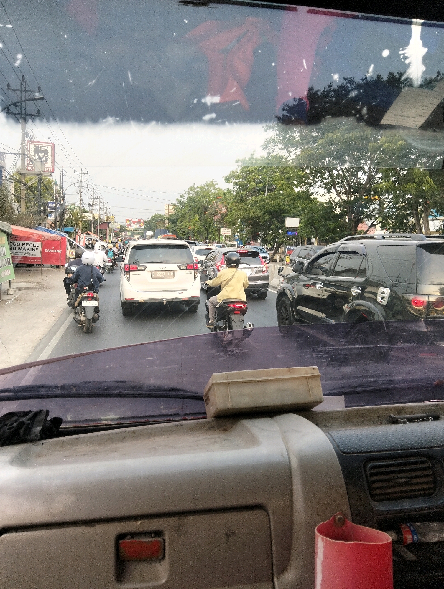 The image depicts a traffic jam on a city street, likely in Semarang, Indonesia, as suggested by the context of the image description. The perspective is from inside a vehicle, looking through the windshield. In the foreground, the dashboard of the vehicle is visible, with a red plastic cup on the right.
The middle ground is dominated by the road and the surrounding vehicles. Several motorcycles are present, with riders wearing helmets and casual clothing. A white SUV, possibly a Toyota Innova, is in the center of the road. To the right, a black SUV, likely a Toyota Fortuner, is also visible. The vehicles are stopped or moving very slowly, indicating heavy traffic.
The background shows buildings, trees, and utility poles with numerous wires, characteristic of an urban environment in Indonesia. Some shops and signs are partially visible along the roadside, with one sign appearing to be for a "Supermarket." The sky is overcast, suggesting a cloudy or hazy day. The overall scene conveys a sense of congestion and the daily commute in a busy Indonesian city.