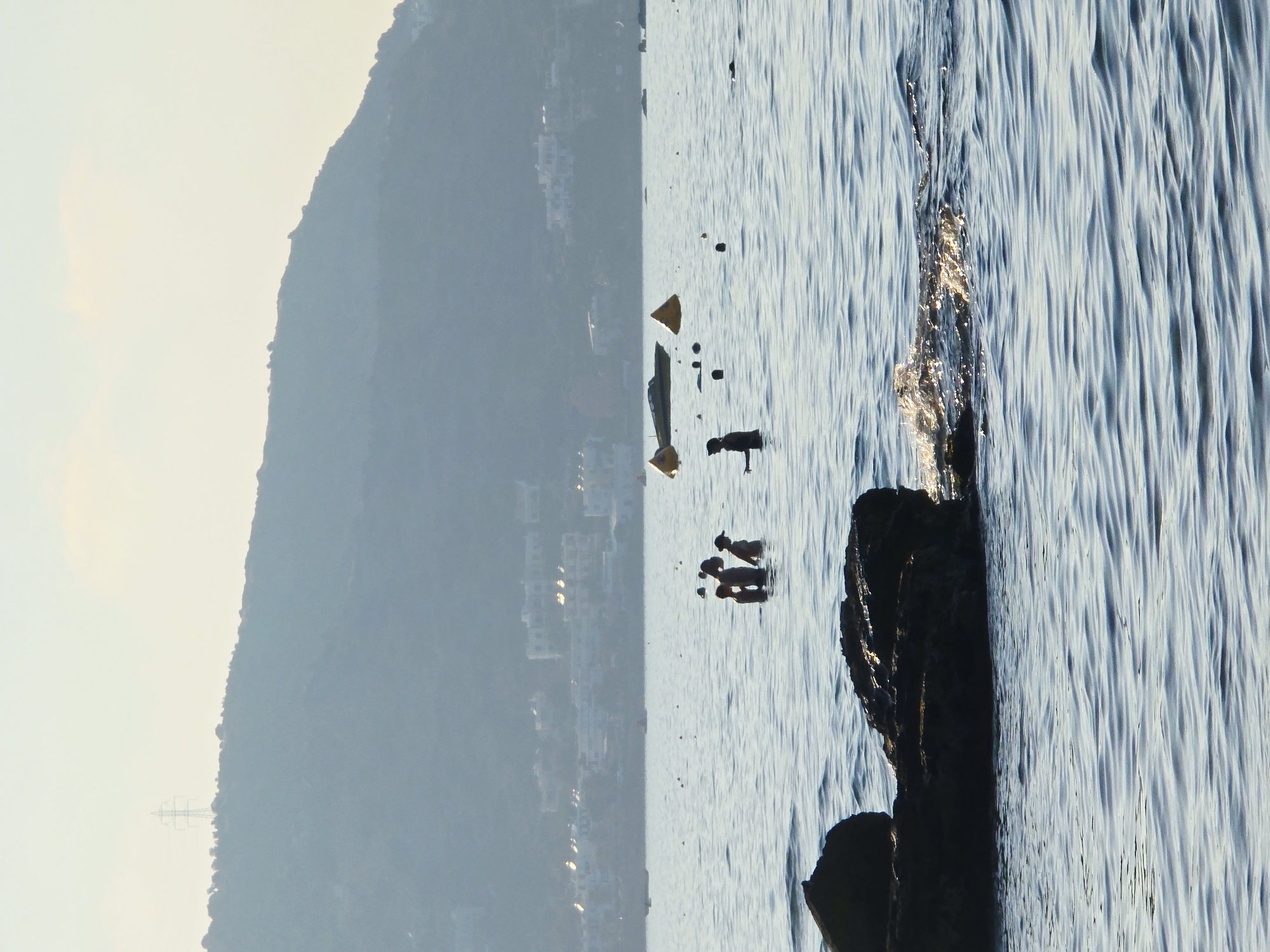 The image depicts a serene coastal scene in Malia, Greece, under a hazy sky. In the foreground, dark, jagged rocks jut out of the tranquil blue water, with sunlight glinting off the ripples. Further out in the sea, several individuals are visible, their silhouettes dark against the water. Some are wading, while others appear to be swimming or playing. In the middle ground, a few buoys and a small boat are anchored, suggesting a designated area for water activities. In the background, a hillside covered with vegetation rises above a cluster of white buildings, indicative of a coastal town. A solitary electricity pylon stands atop the hill, a testament to the area's infrastructure. The overall impression is one of a peaceful summer day at the beach.