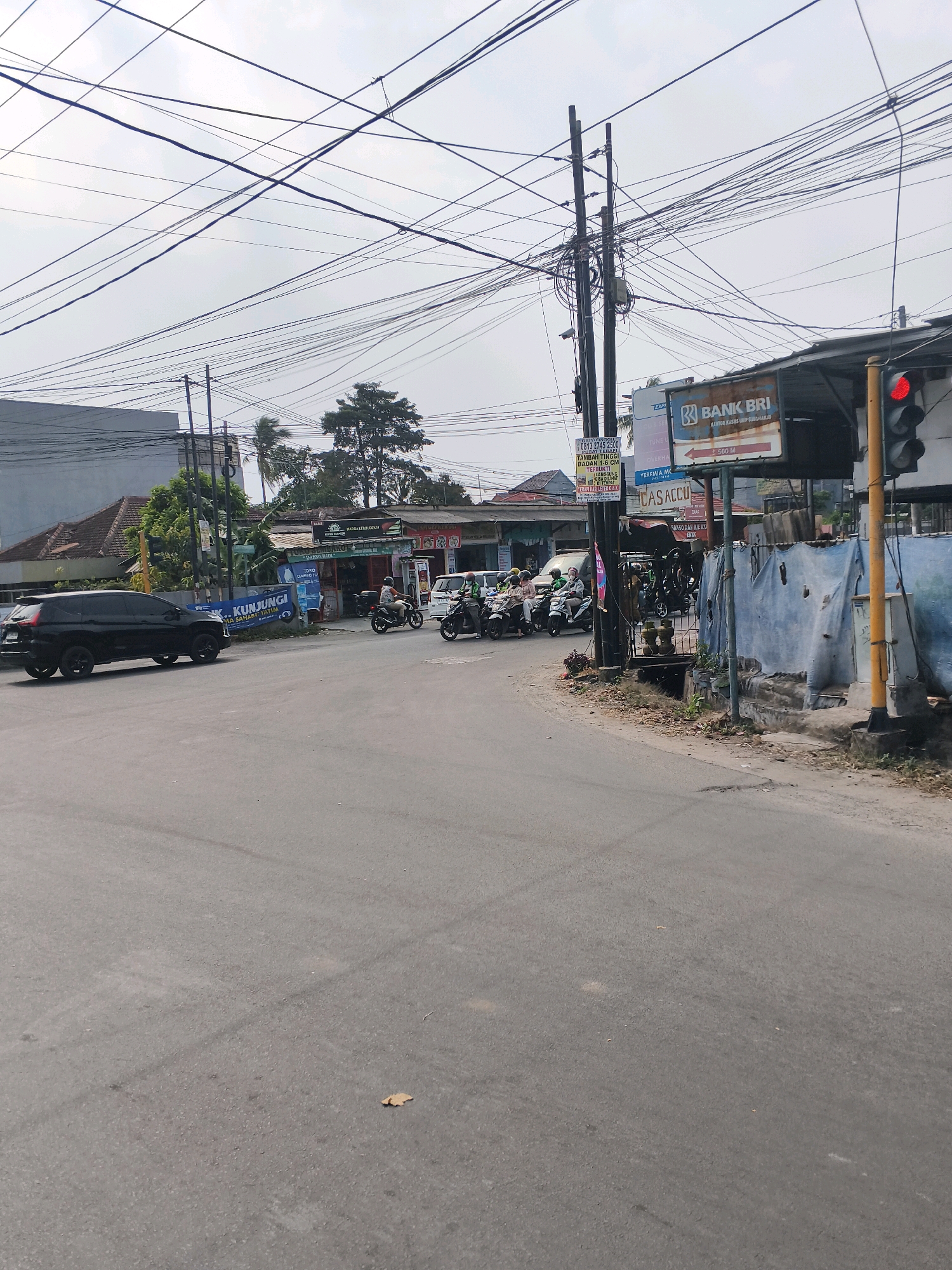 The image shows a street scene in Bandar Lampung, Indonesia. A black SUV is parked on the left side of the road. Several motorcycles, some with riders, are stopped at an intersection. A traffic light is visible and is red, indicating that traffic should stop. A sign for "BANK BRI" is prominent on the right side of the intersection. The sky is overcast, and there are many power lines visible overhead. The area appears to be a commercial or mixed-use district with buildings and shops lining the street.